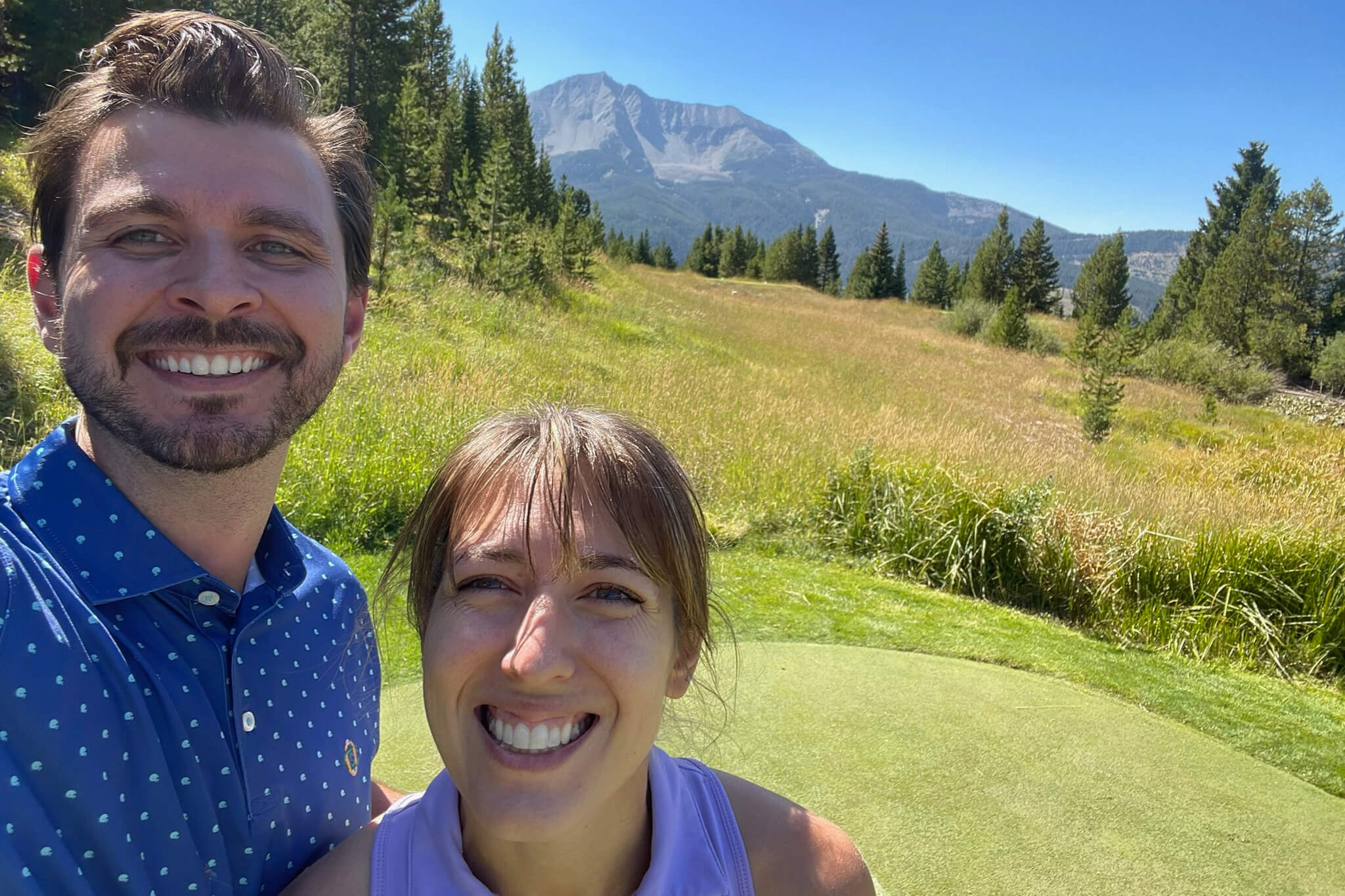 Smiling man and woman take a sunny selfie in a grassy meadow with pine trees and a mountain peak in the background.