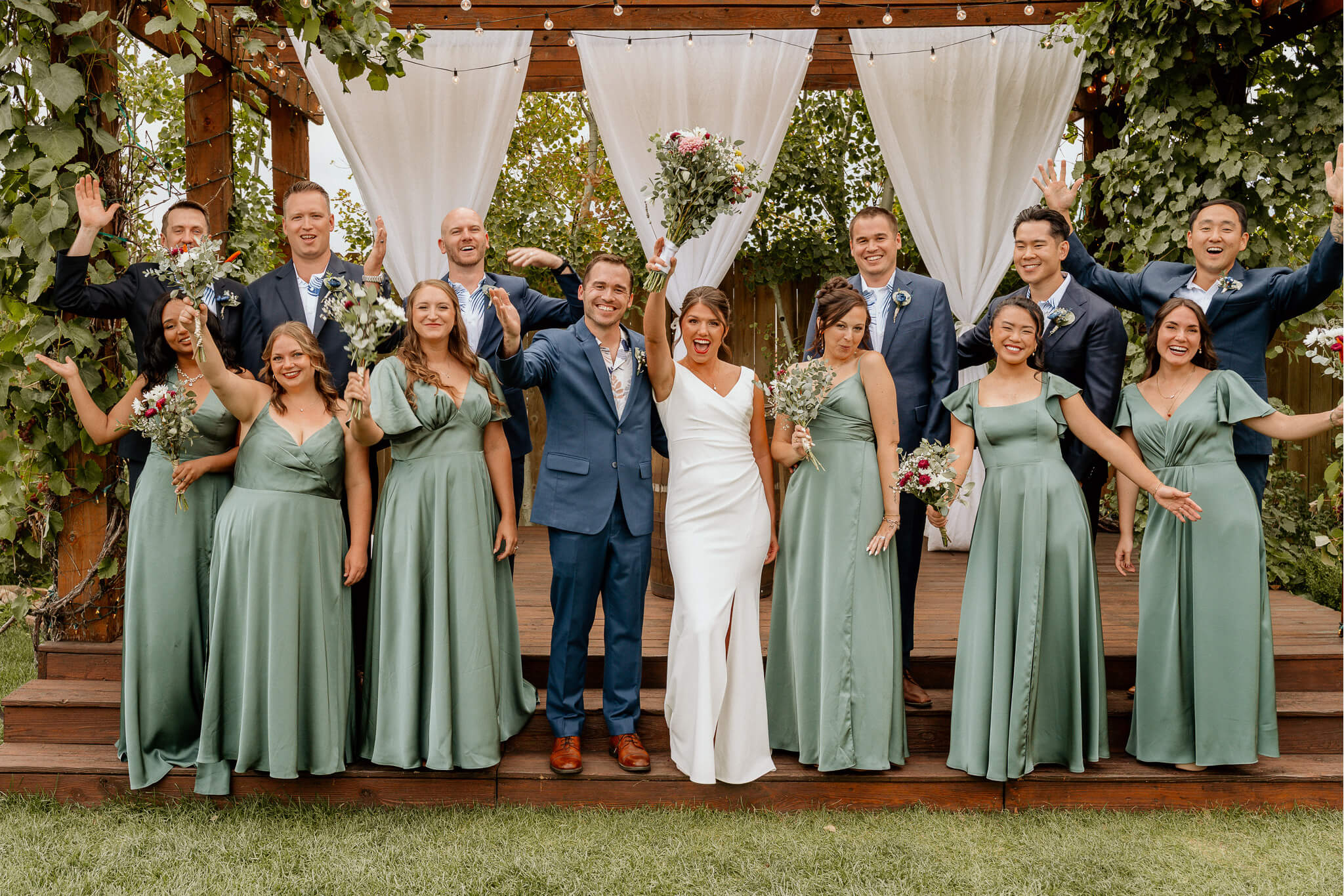 Bride and groom with wedding party outdoors, bridesmaids in sage gowns and groomsmen in navy suits, raising bouquets and hands in celebration under a wooden arch.