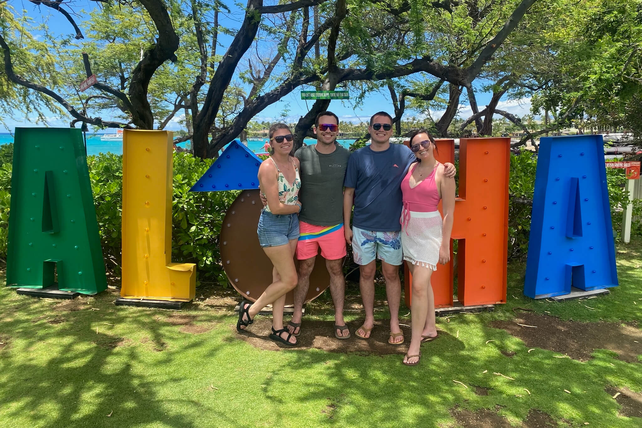 Group of four friends posing smiling in front of large colorful letters spelling 'ALOHA' at a tropical beachside park. Behind them are green trees and blue ocean under a sunny sky.