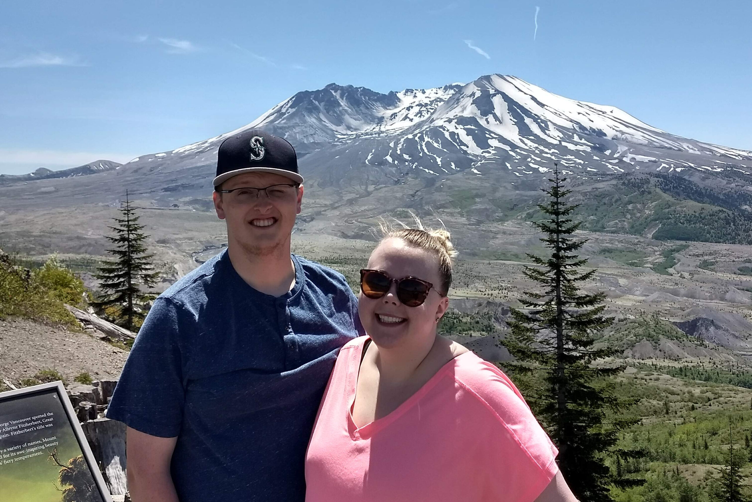 Smiling couple taking a selfie in front of a snow-capped mountain and clear blue sky, with trees in the foreground.