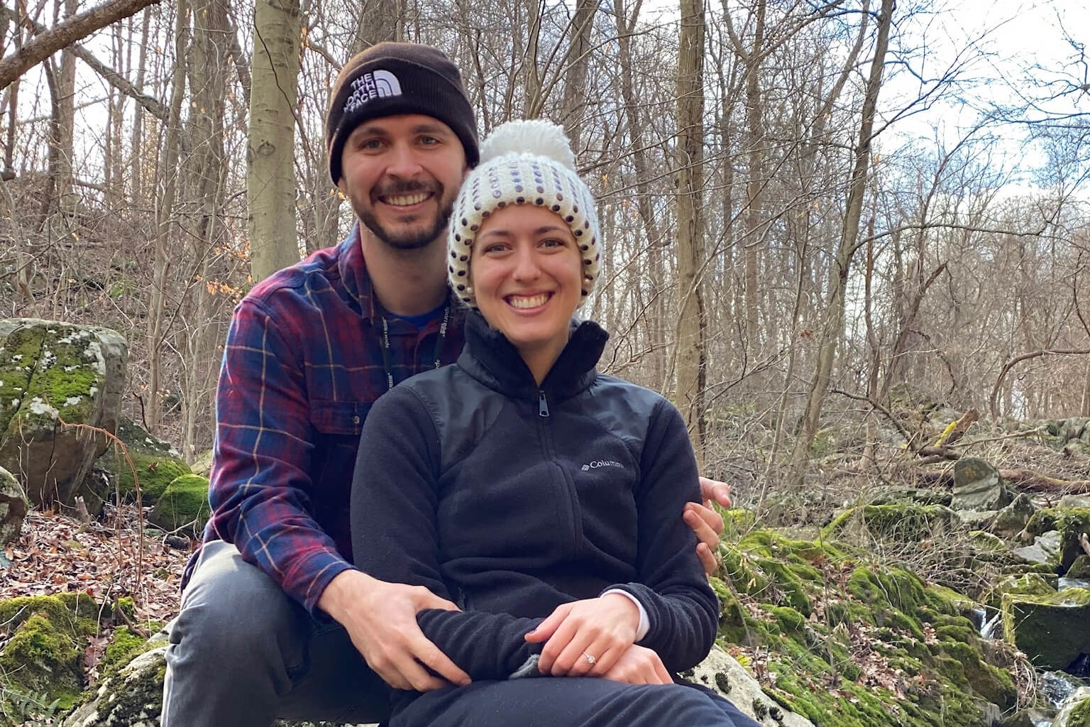 Smiling couple sitting on mossy rocks in a bare-wooded forest, embracing outdoors in winter.