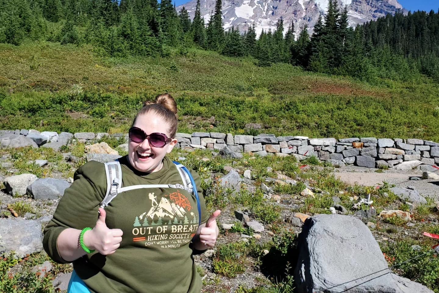 Smiling woman in sunglasses gives two thumbs up on a rocky hiking trail with a stone wall and forested mountains behind her.