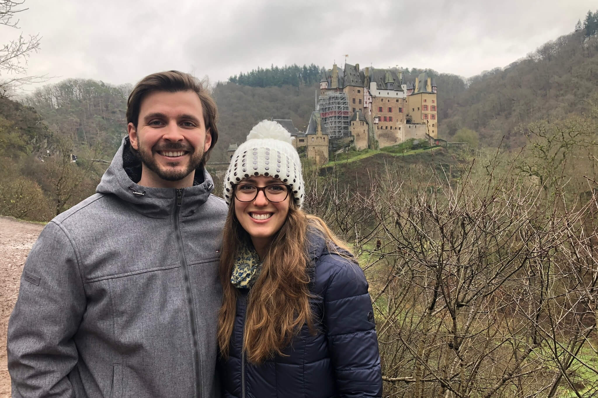 Smiling couple posing outdoors with a medieval castle in the cloudy background and bare vines in the foreground.