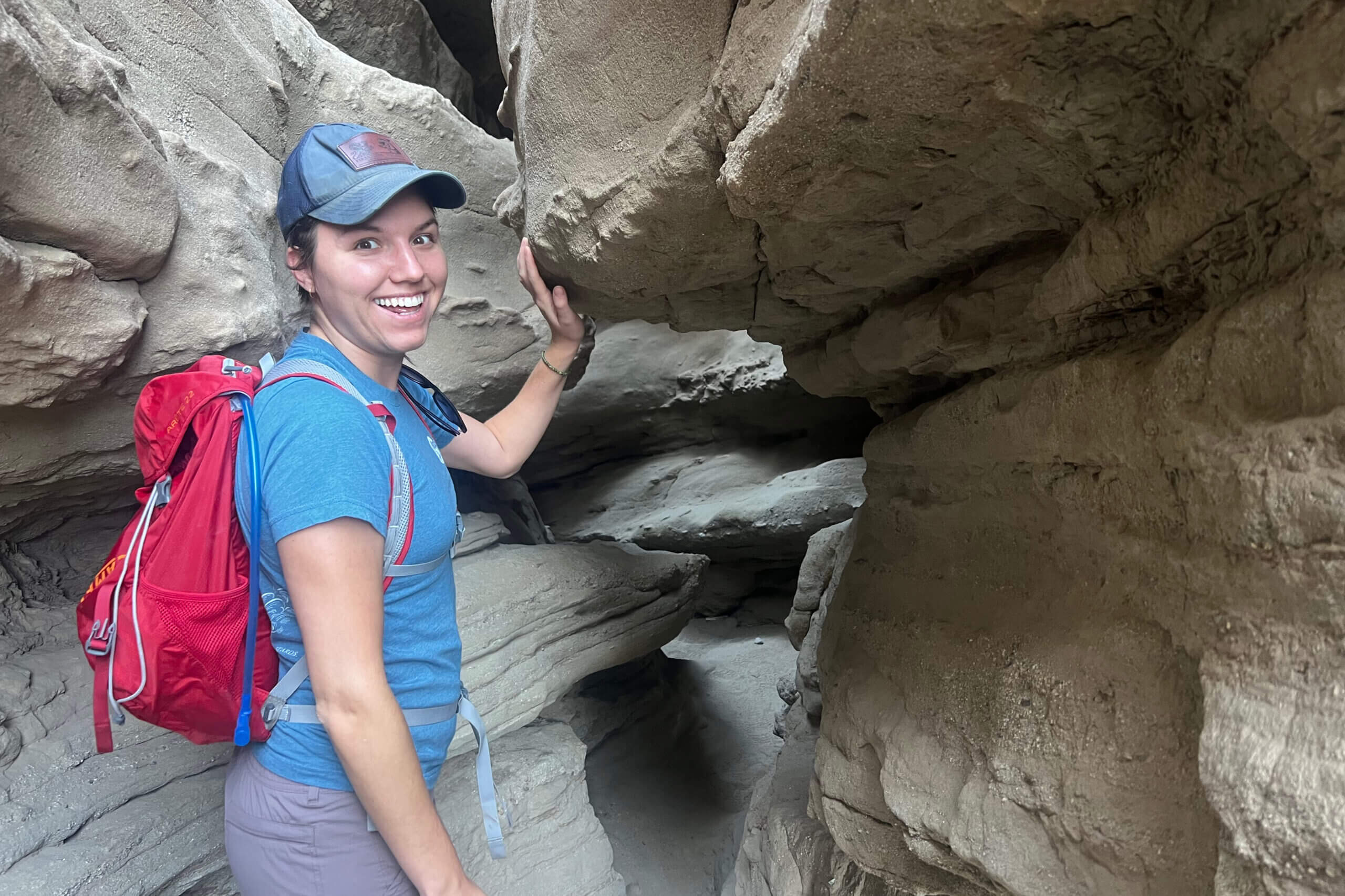 Woman wearing a blue shirt and red backpack squeezes through a narrow rocky crevice in a canyon-like formation.