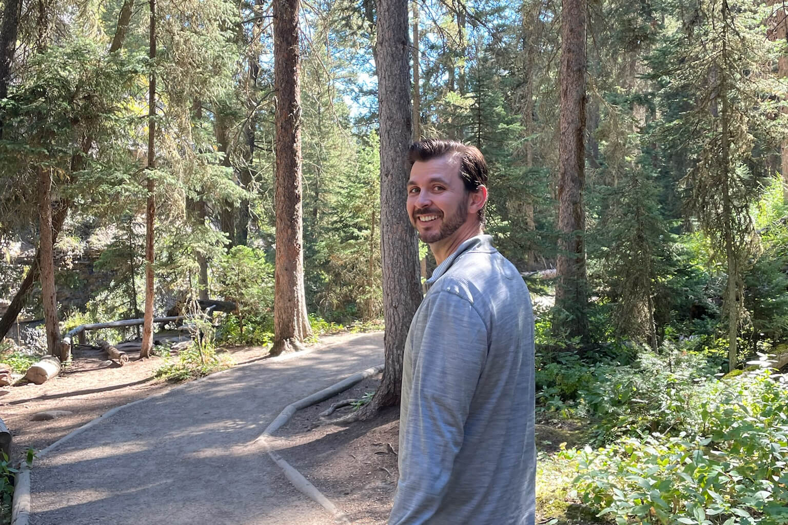 Man in a light-blue jacket smiles at the camera on a forest path lined with tall trees.