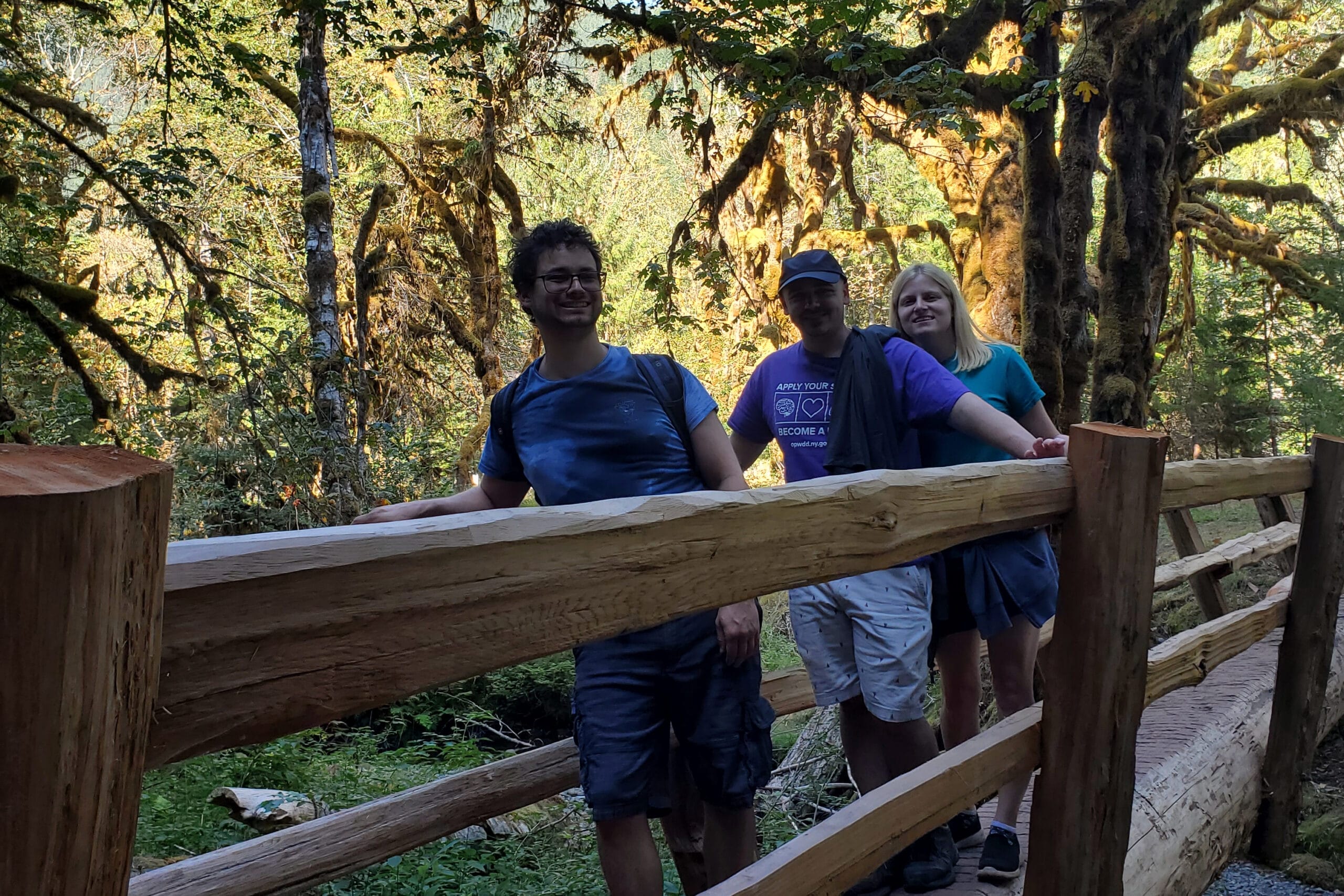 Three friends pose along a mossy wooden railing in a sunlit forest, smiling at the camera.