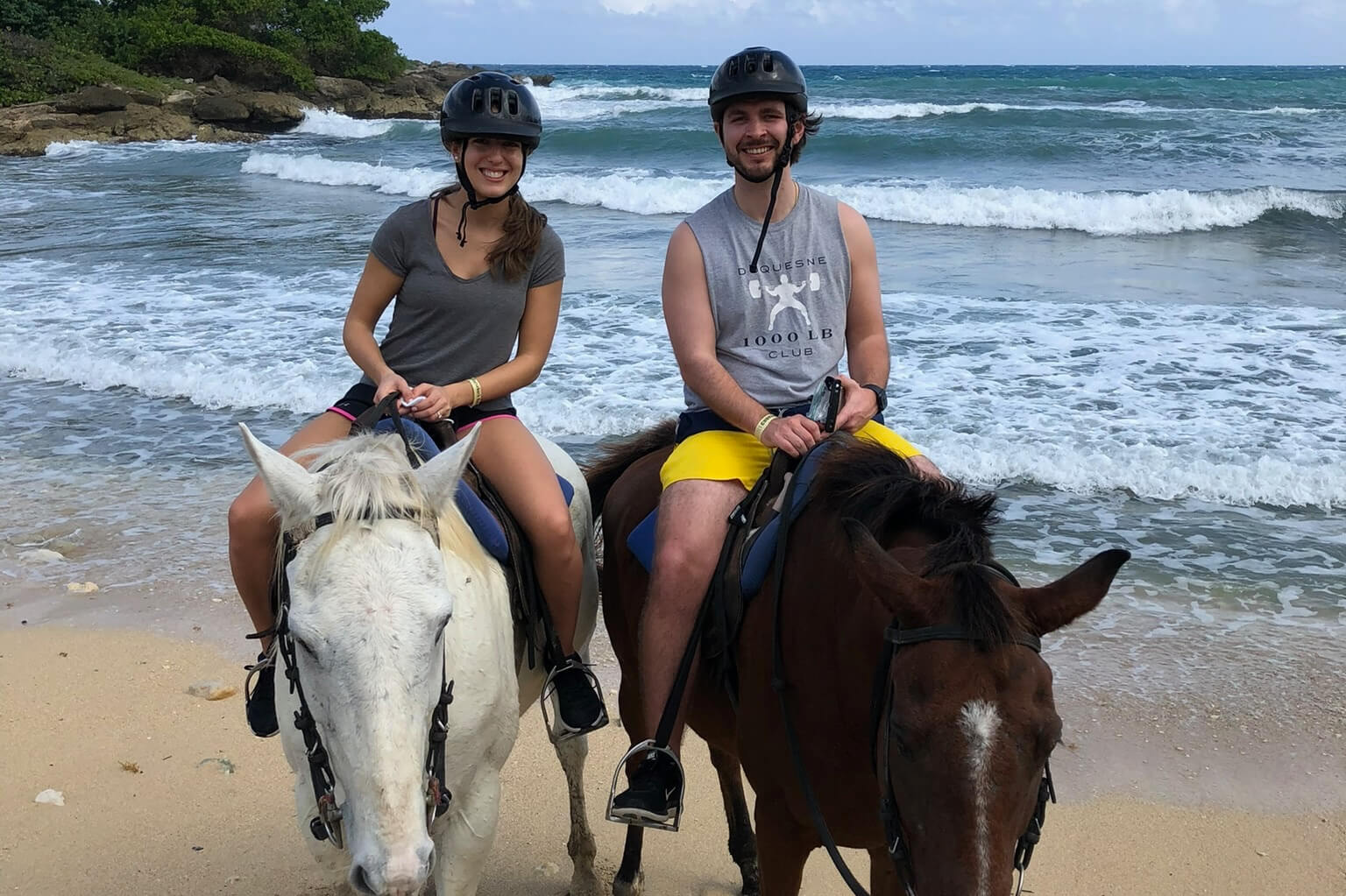 Two riders in helmets ride white and brown horses along a sandy beach with waves in the background.