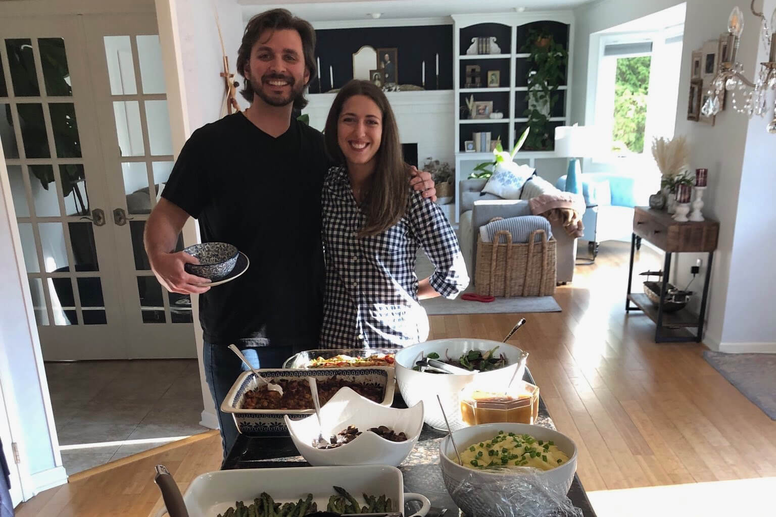 Couple stands at a kitchen island with bowls of food, smiling at the camera in a bright living room.