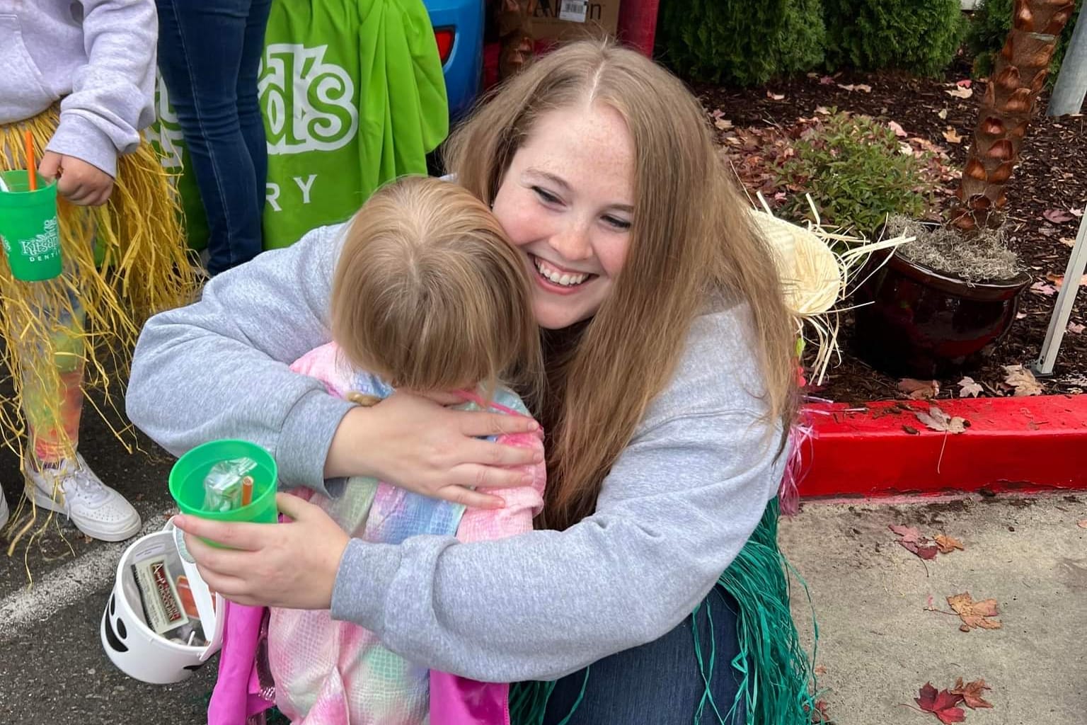 Smiling young woman in a gray hoodie hugs a blonde child outdoors at a festival, with a green cup in the child's hand.