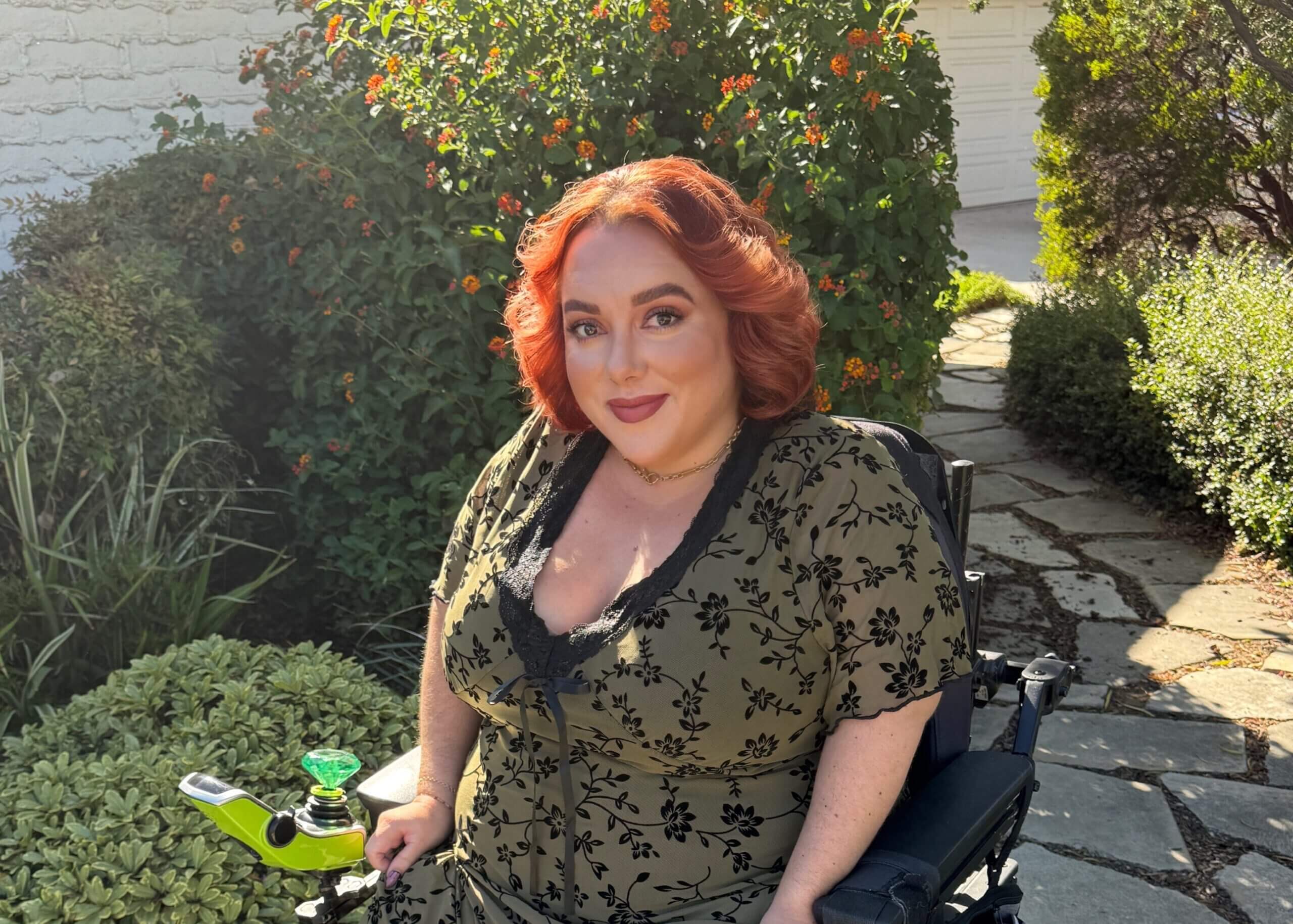 Woman with red hair in a floral beige dress sits in a wheelchair in a sunny garden.
