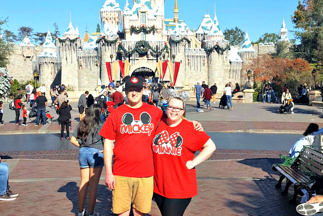 Two friends in red Mickey and Minnie shirts pose arm-in-arm in front of a snow‑topped castle at a Disney park.”,