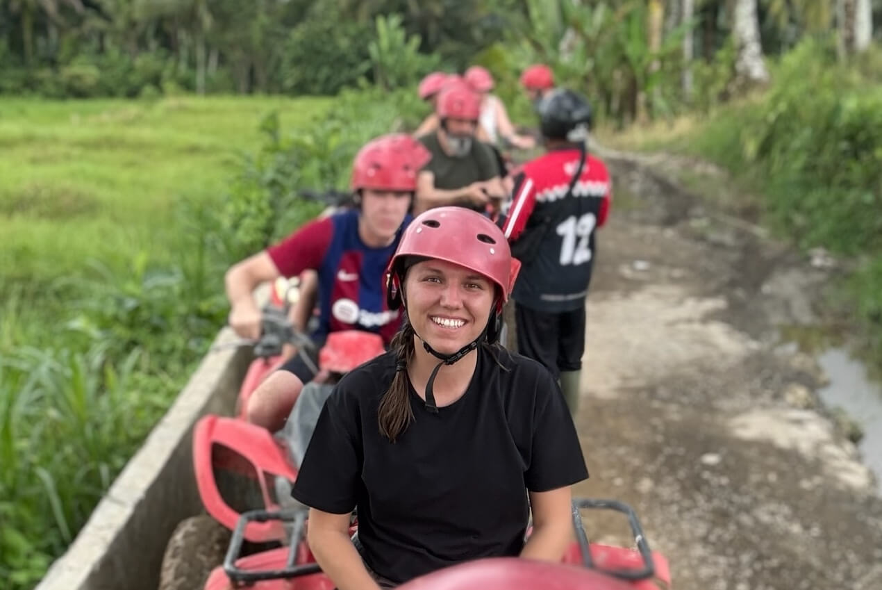 Smiling woman in a red helmet and black shirt in the foreground during an outdoor group activity, with teammates in red helmets behind her on a dirt path.