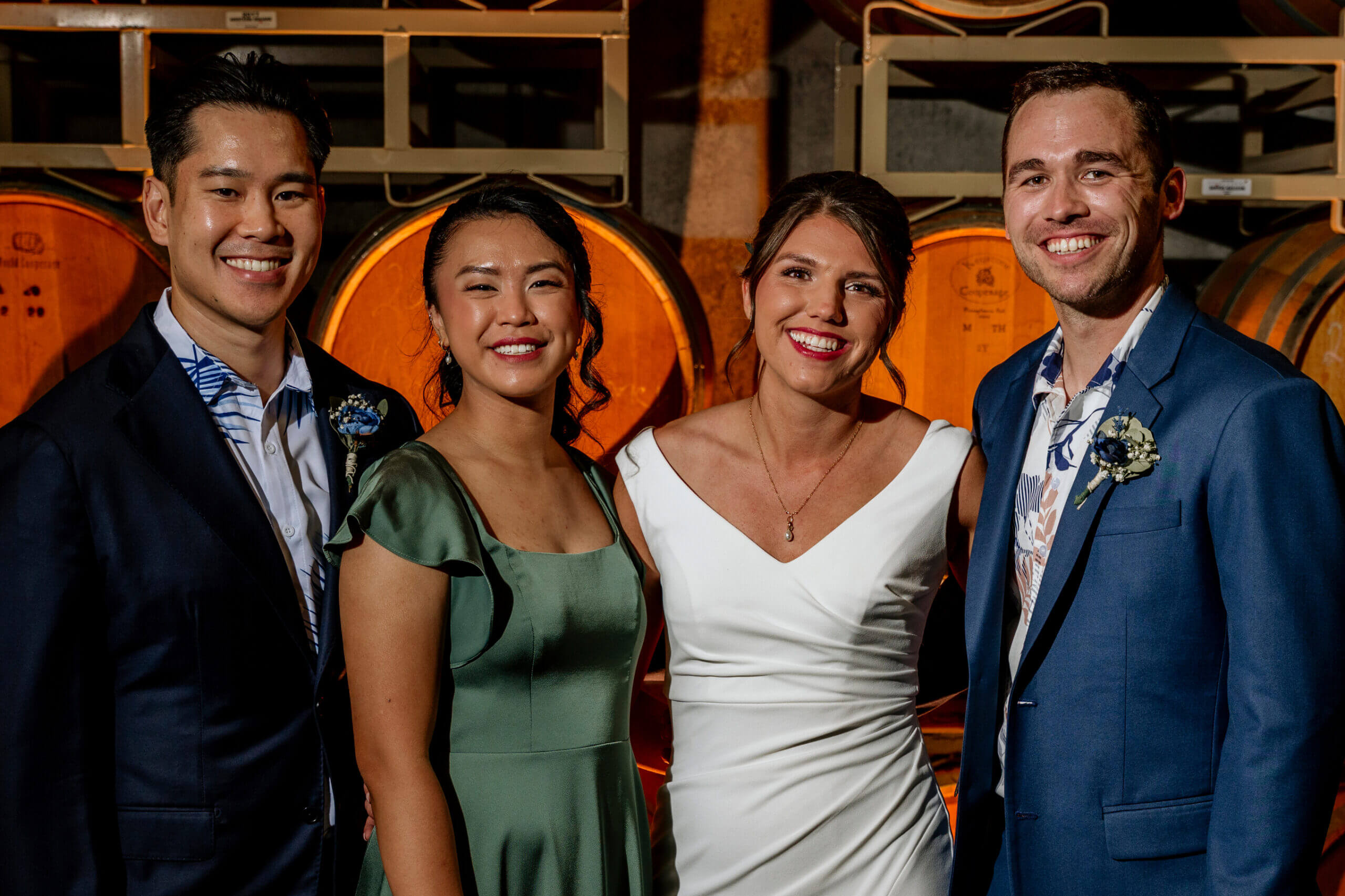 Four friends in formal attire posing together in a winery with wooden barrels behind them, smiling at the camera.