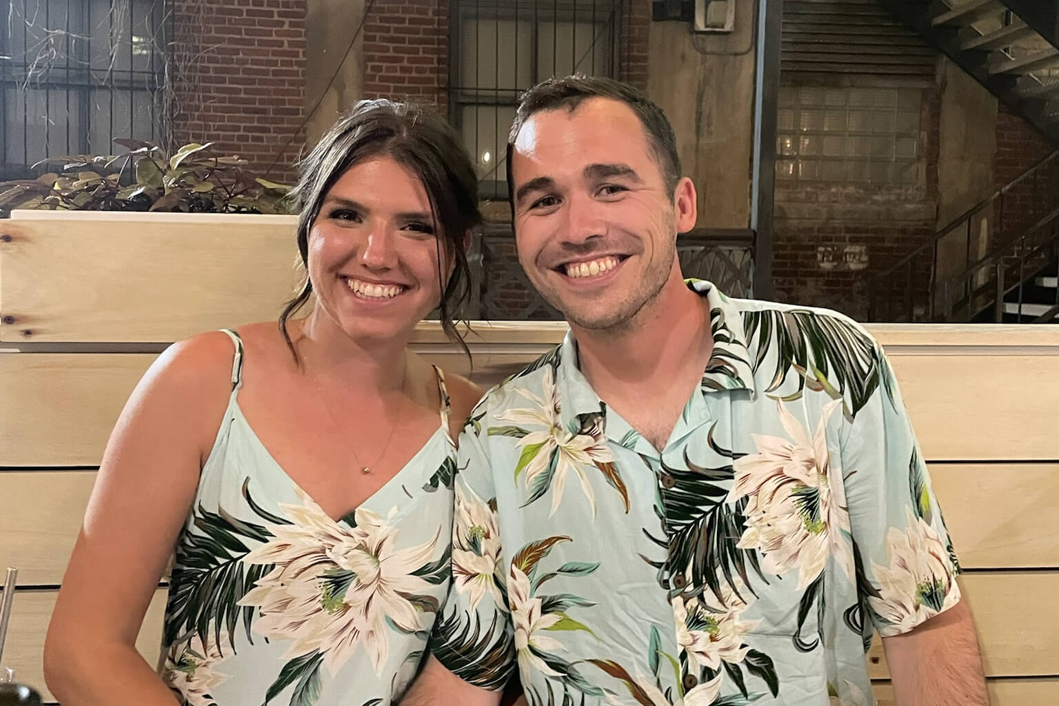 Smiling couple in matching floral shirts seated at a booth in a brick-walled restaurant.