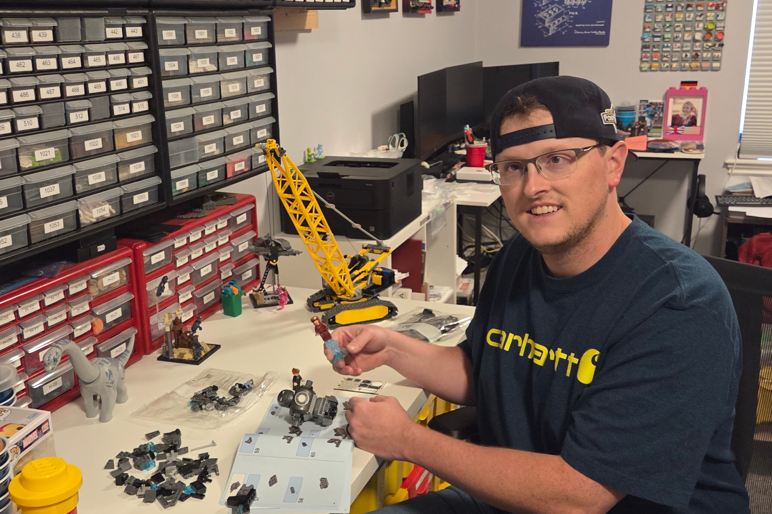 Man wearing a backward cap builds a yellow LEGO crane at a cluttered workbench, instruction booklet open nearby.