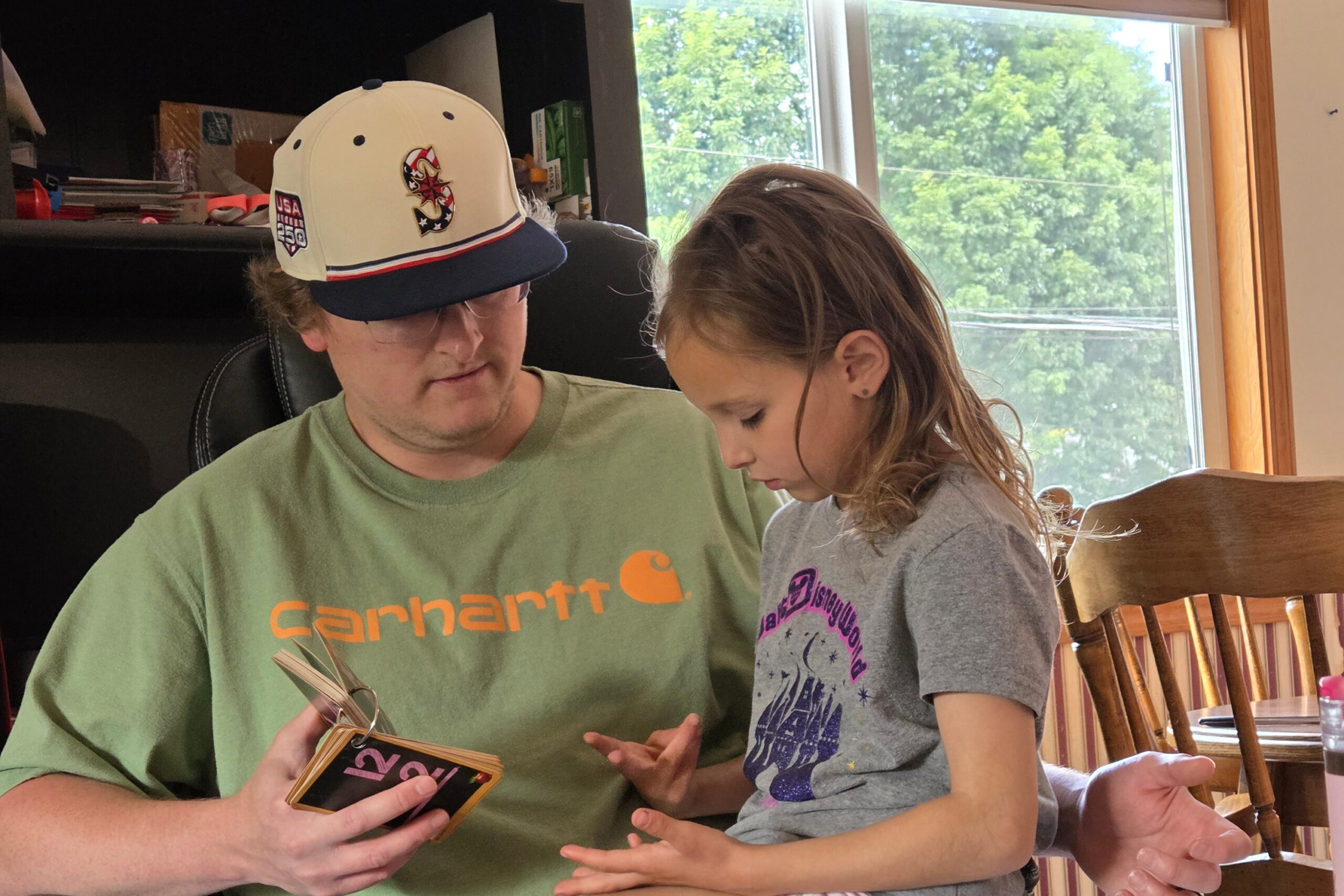 Man in a green Carhartt shirt wearing a white cap with patches, showing a small device to a girl as they sit at a table by a window.
