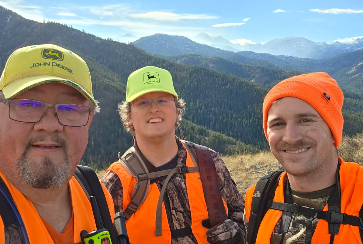 Three men wearing bright orange safety vests and John Deere hats pose on a mountain ridge with a forested valley behind them.