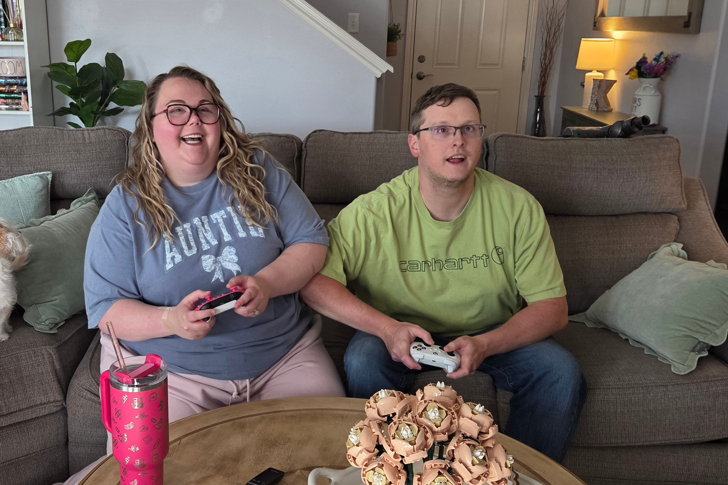 Two people sit on a couch and play video games together; the woman on the left laughs while the man on the right concentrates on the game.