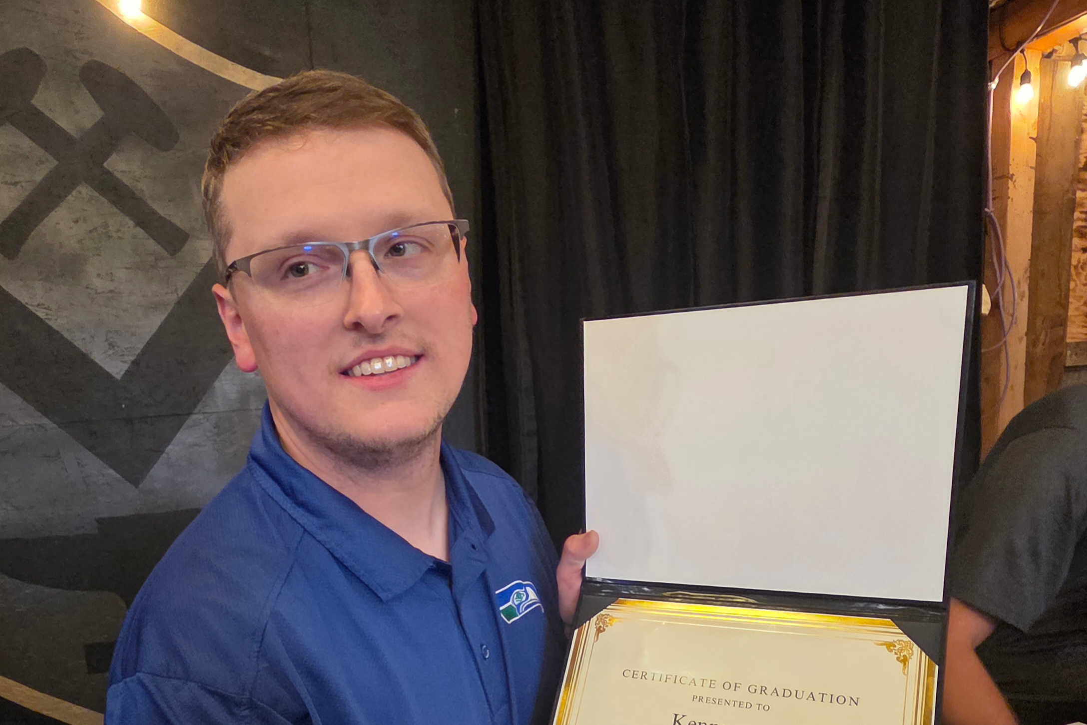 Man in blue shirt smiling while holding a certificate of graduation against a dark backdrop.