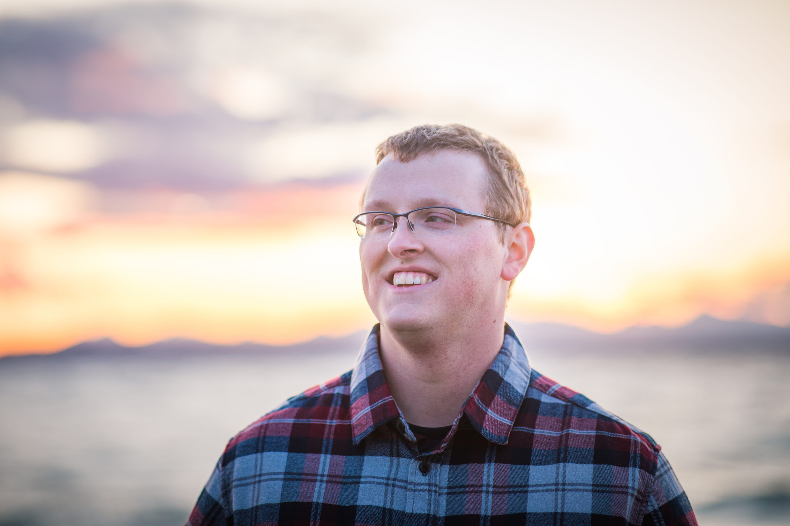 Smiling man with glasses wearing a plaid shirt, sunset over a blurred waterfront behind him.