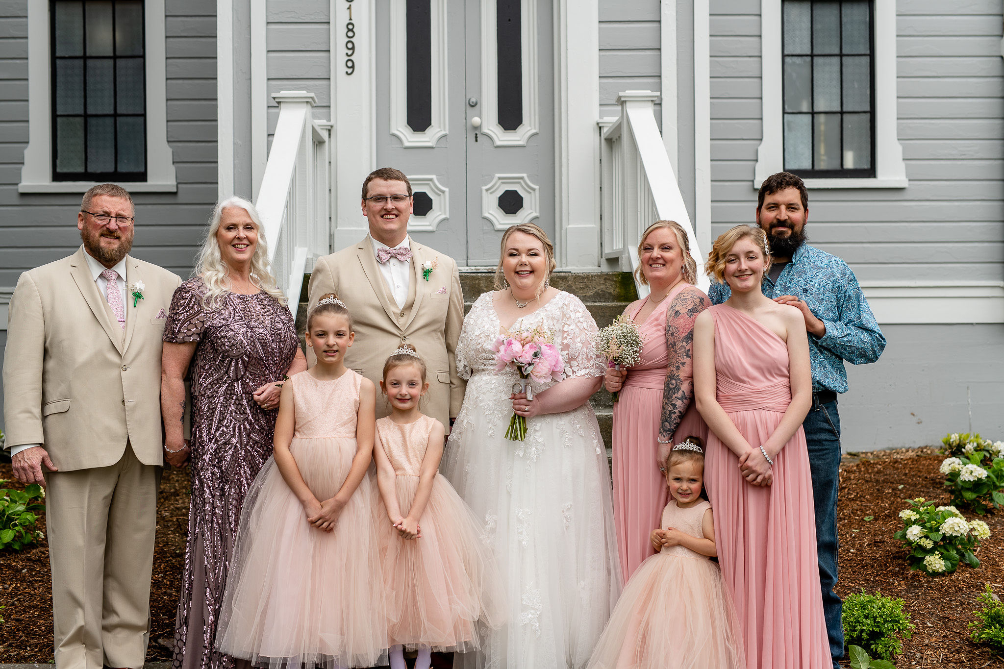 Bride in a white lace gown holds a bouquet, flanked by wedding guests on the front steps of a gray house; bridesmaids in pink dresses and men in beige suits visible in the group.