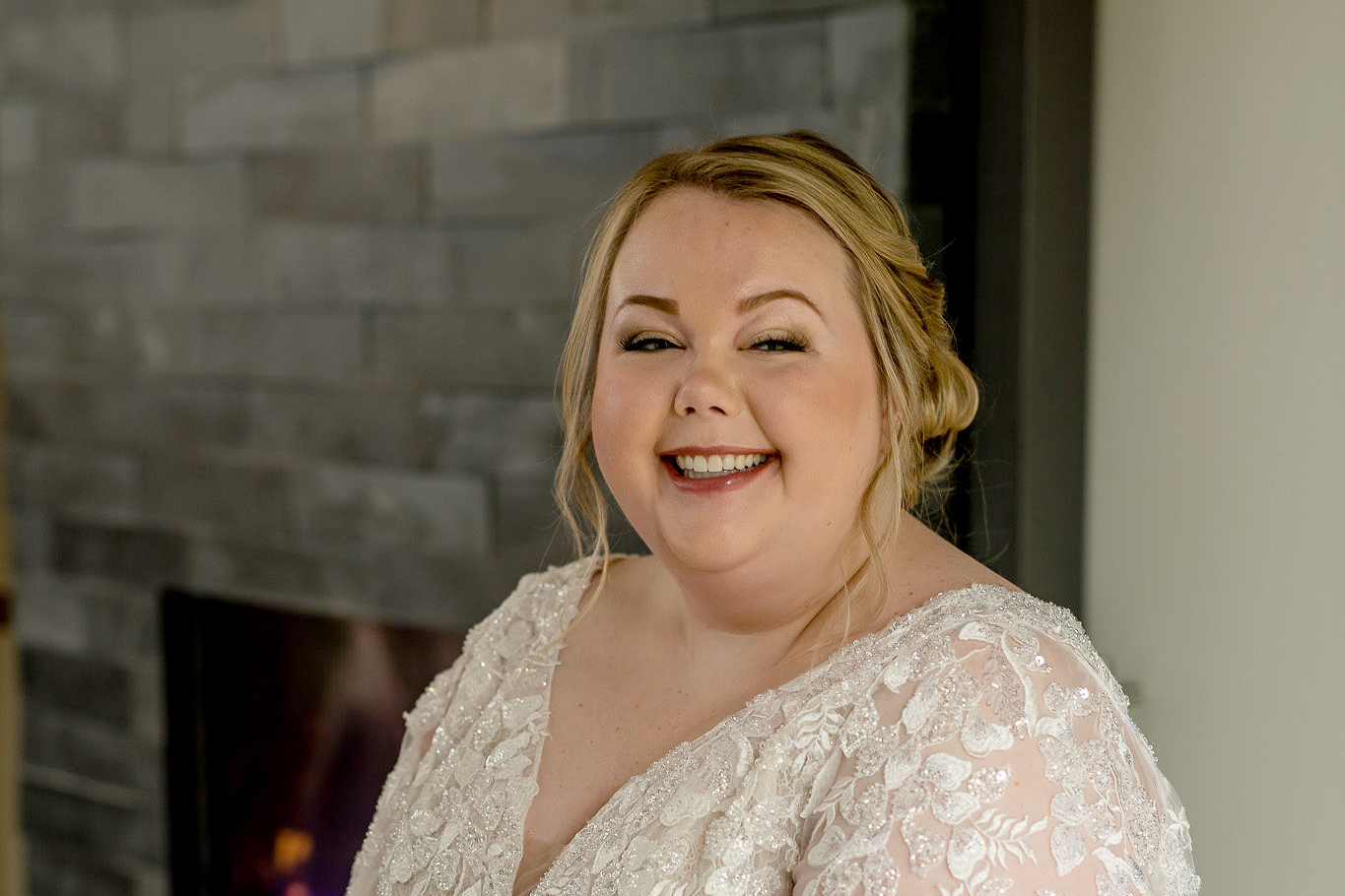 Bride in a beaded lace wedding dress smiling at the camera indoors near a stone fireplace.