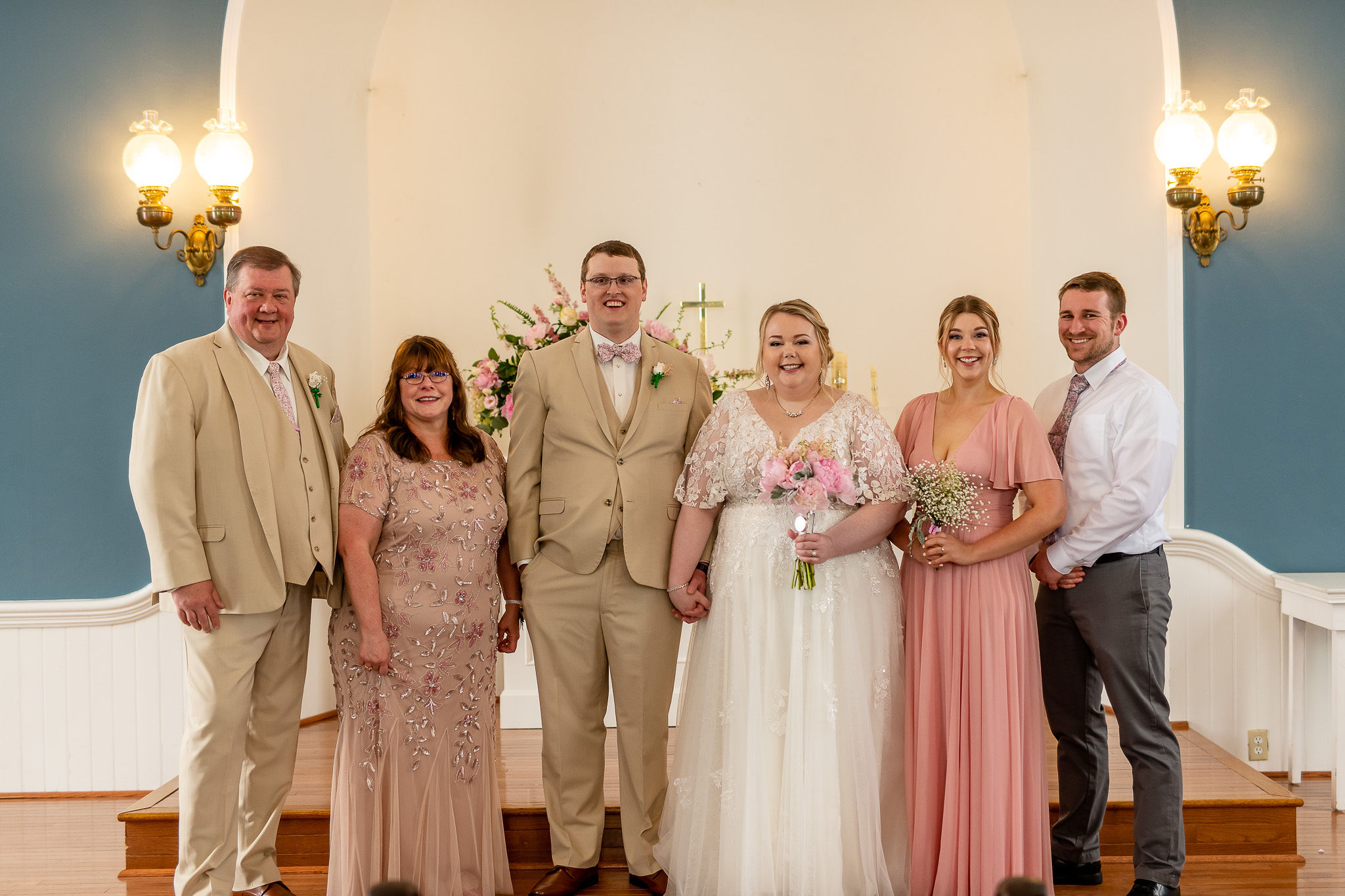 Wedding party posed in front of altar: bride in white lace gown with bouquet, groom in beige suit, bridesmaids in blush dresses, and groomsman in white shirt and tie set against blue walls.
