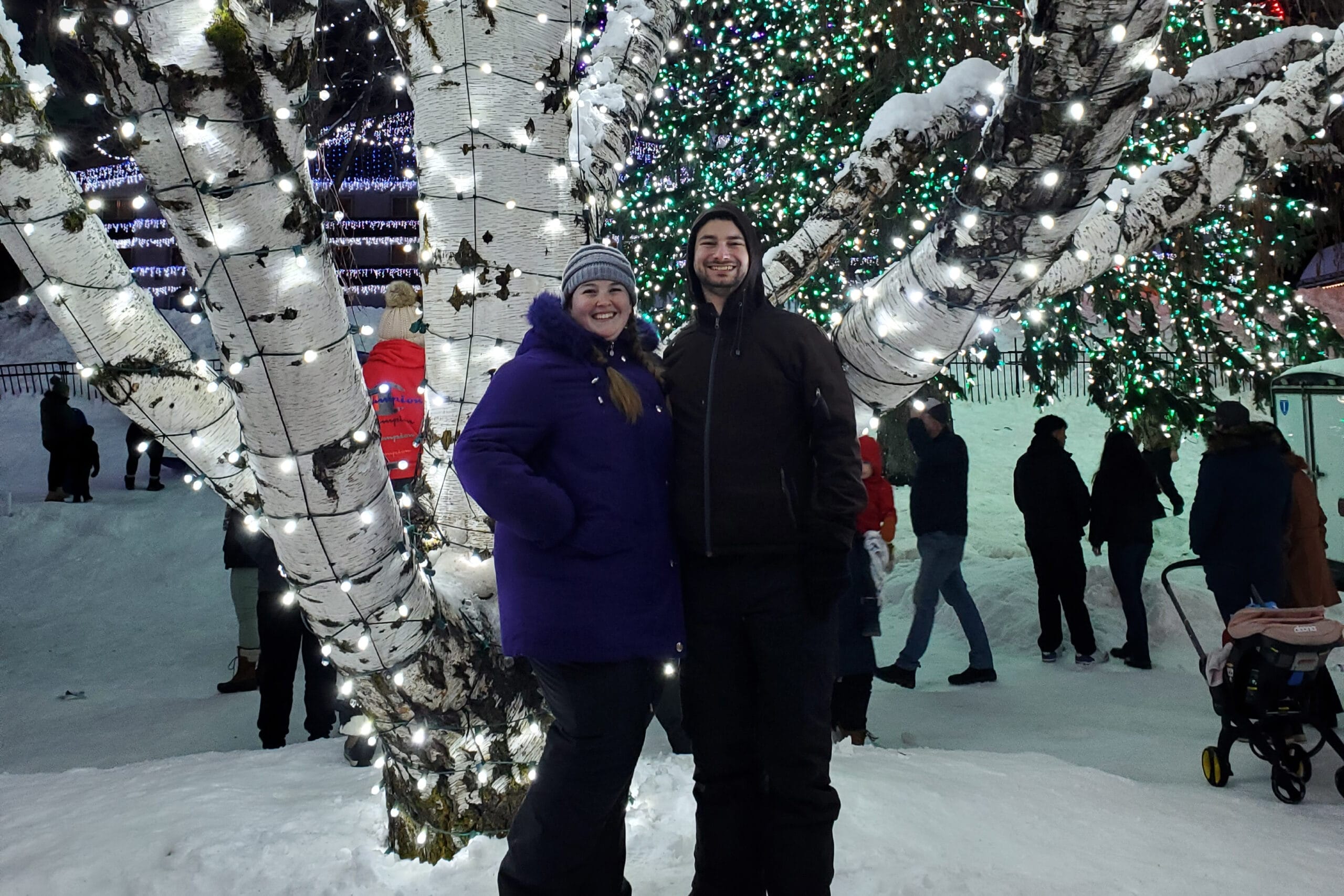 Smiling man and woman in winter coats pose under a tree wrapped in white and green fairy lights in a snowy park at night.