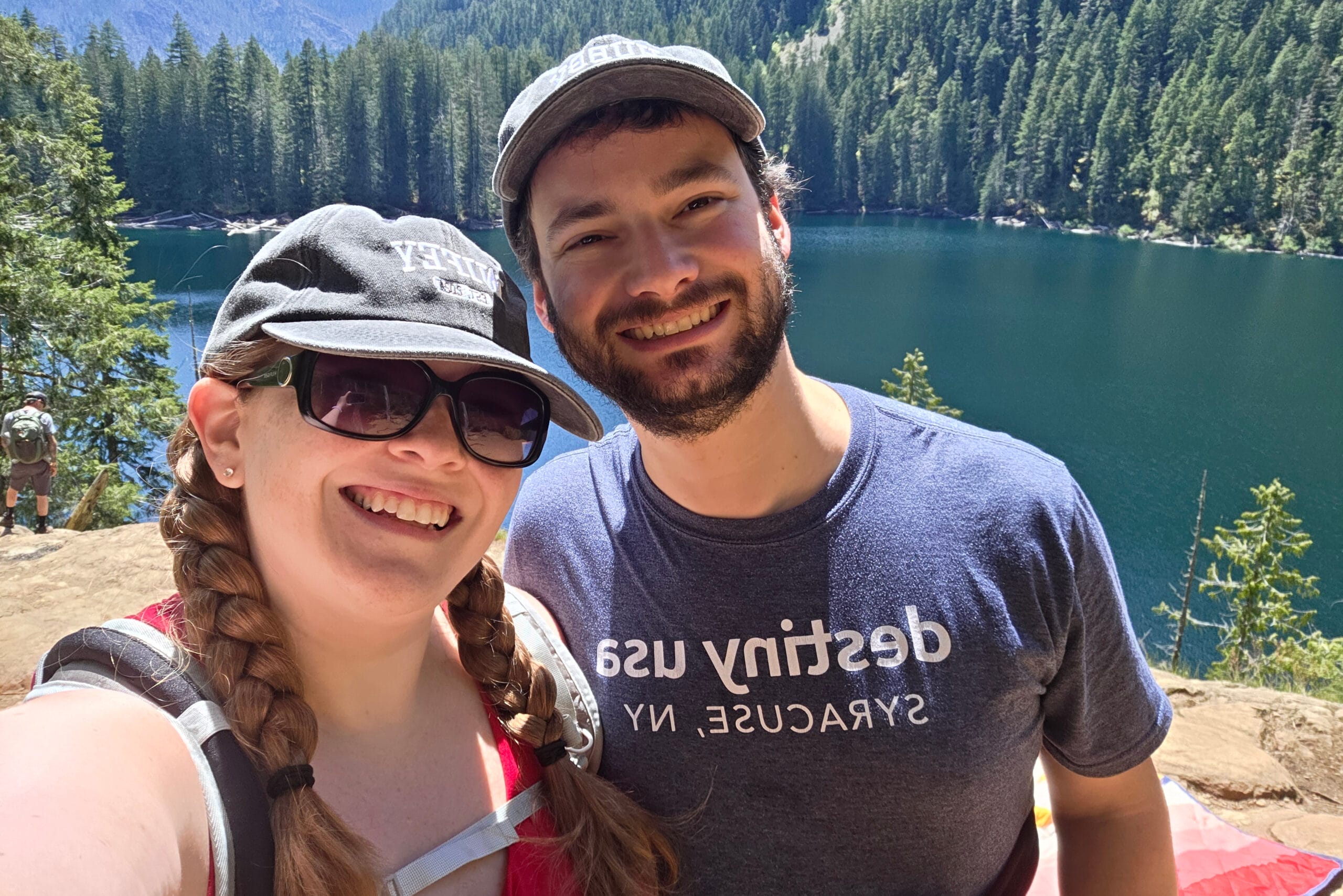 Smiling couple taking a selfie at a forested lakeside with clear blue water for the background.