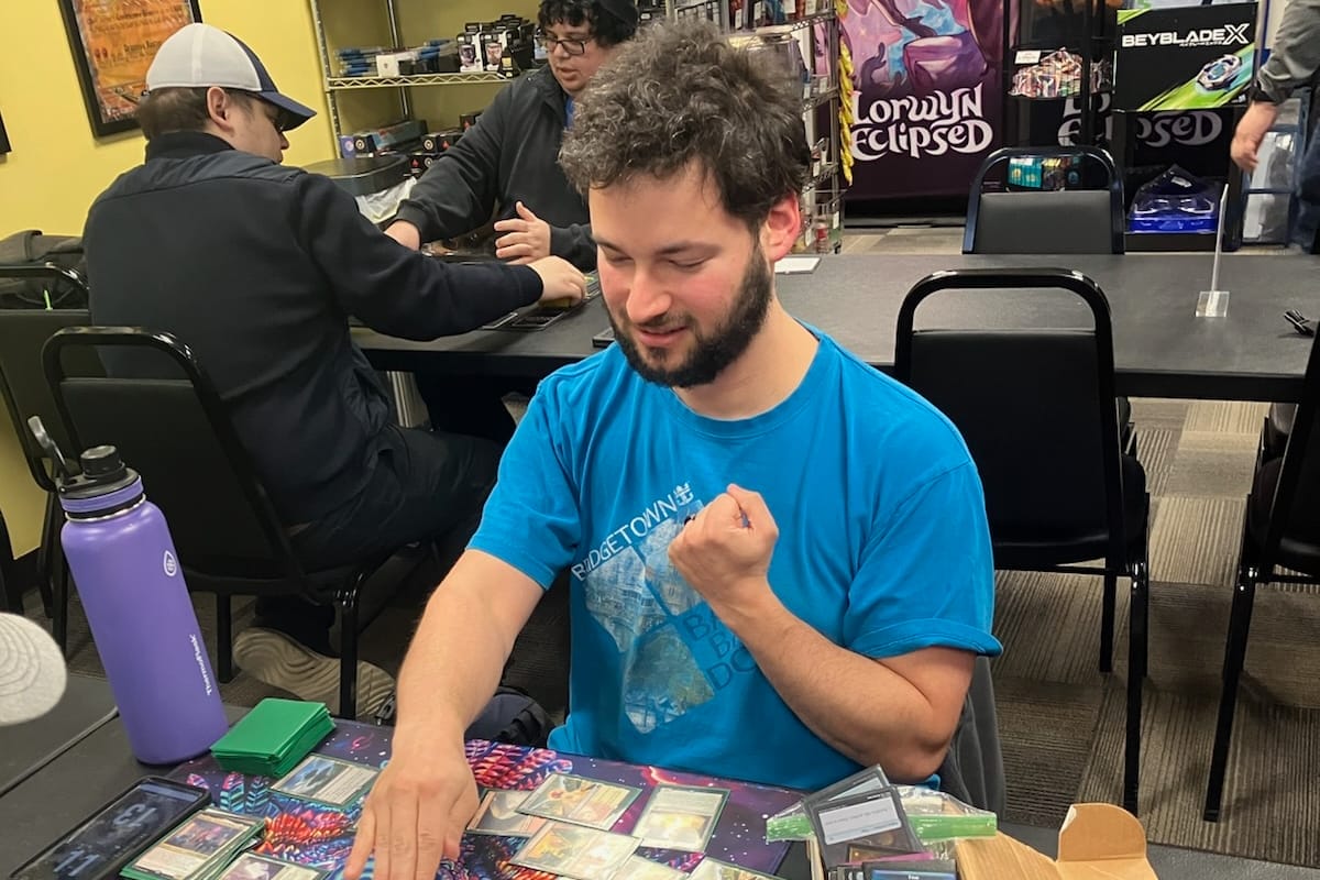 Man in a blue shirt at a tabletop card game, fist raised in celebration; others play in the background in a store.