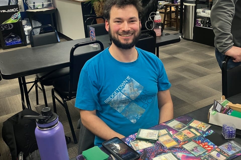 Man with a blue Bridgetown tee sits at a table playing a card game, smiling at the camera in a store.