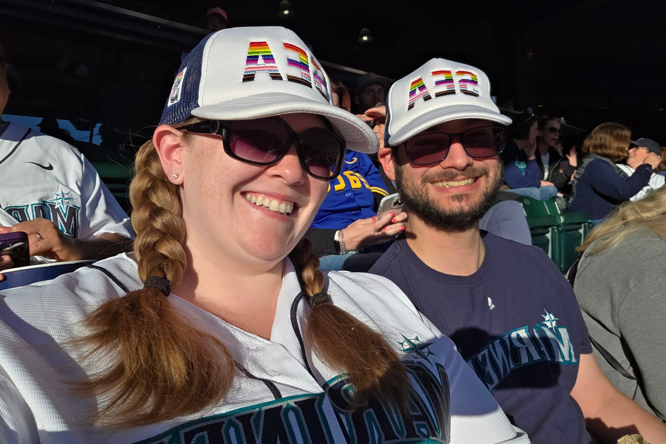 Two fans in sunglasses in a stadium, wearing white caps with rainbow letters and smiling at the camera.