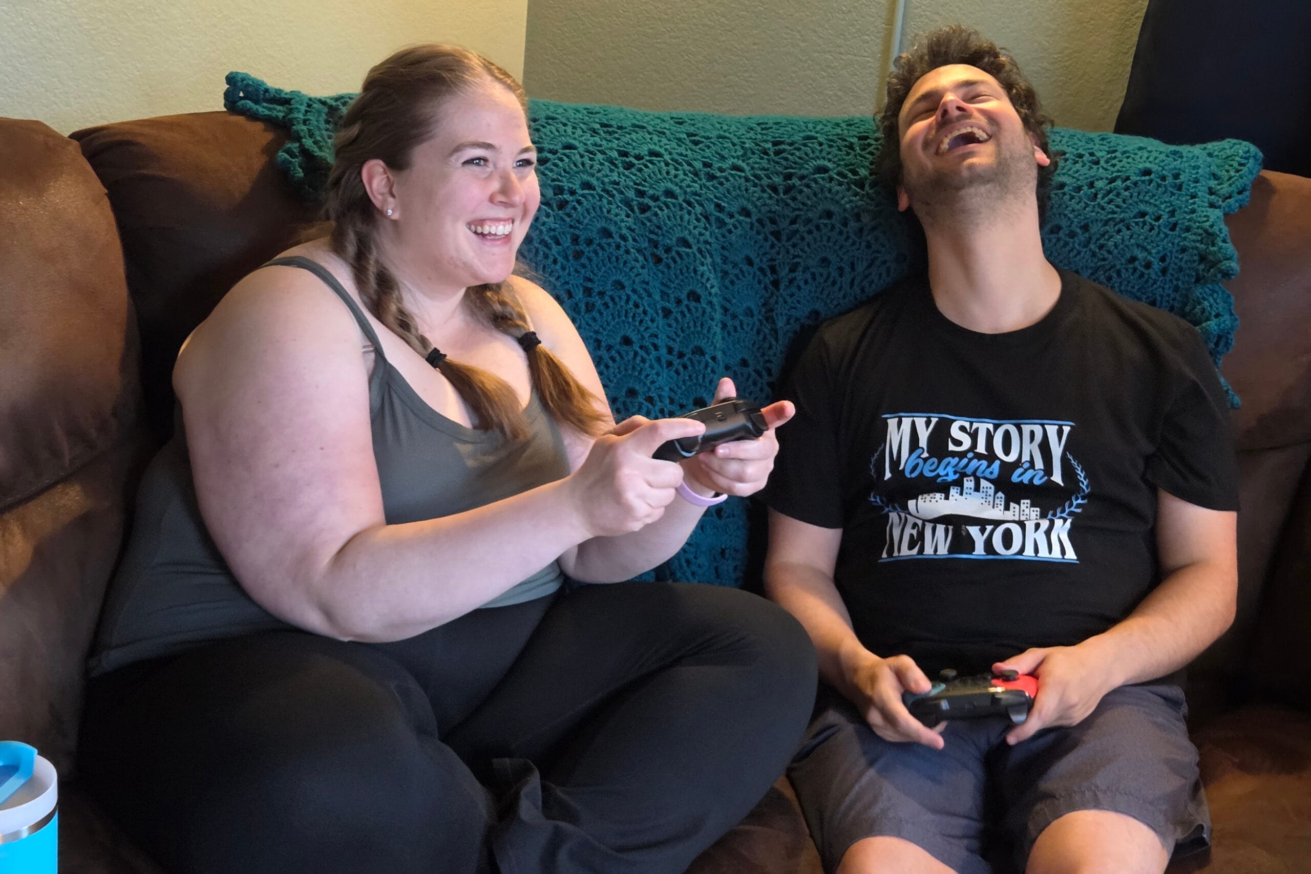 Two adults sit on a couch, smiling and playing video games with controllers in their hands behind a teal crocheted blanket.