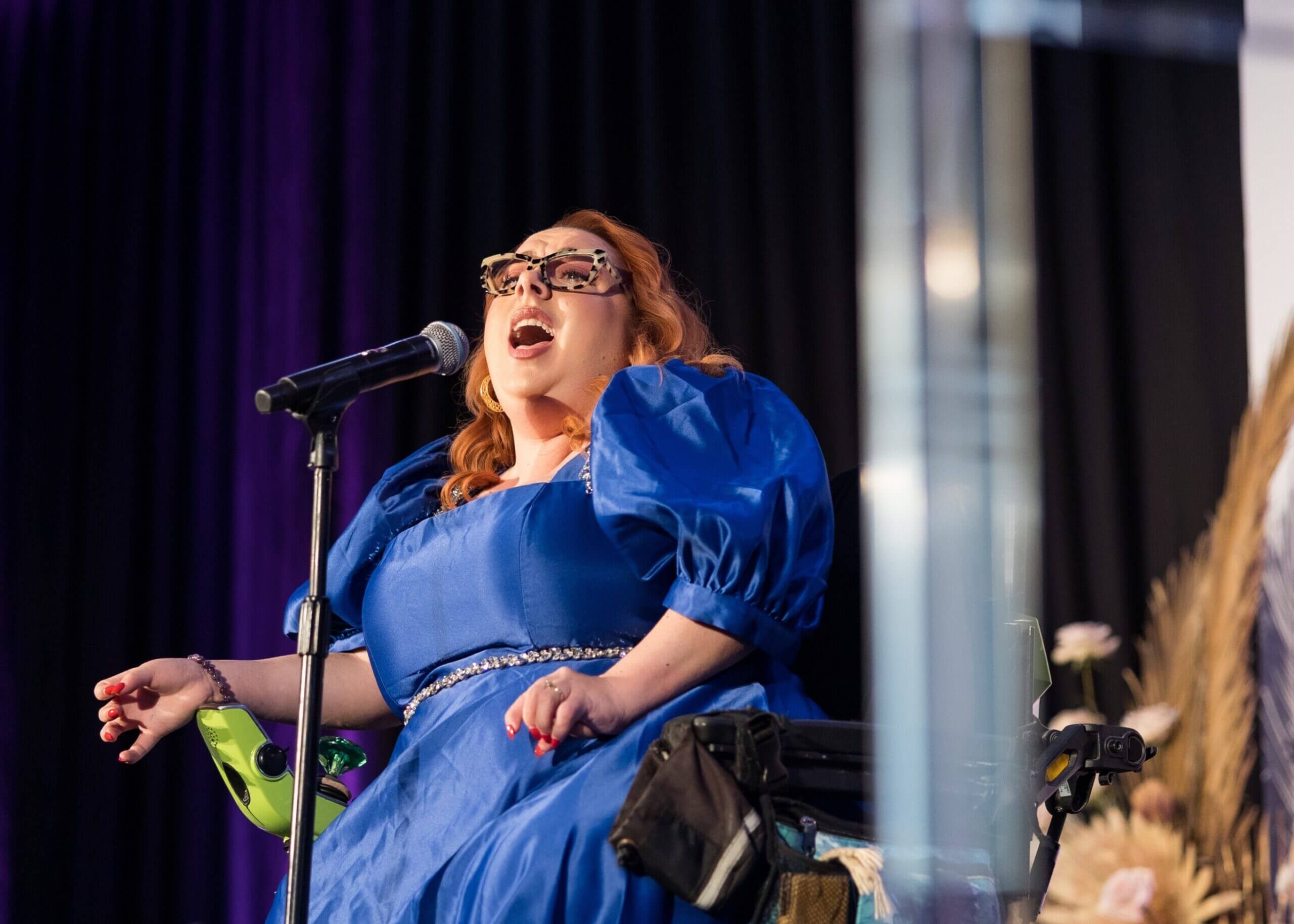 Woman in a royal blue dress sings into a microphone on stage, wearing patterned glasses and a glamorous updo; she appears to be seated in a wheelchair with stage equipment nearby.