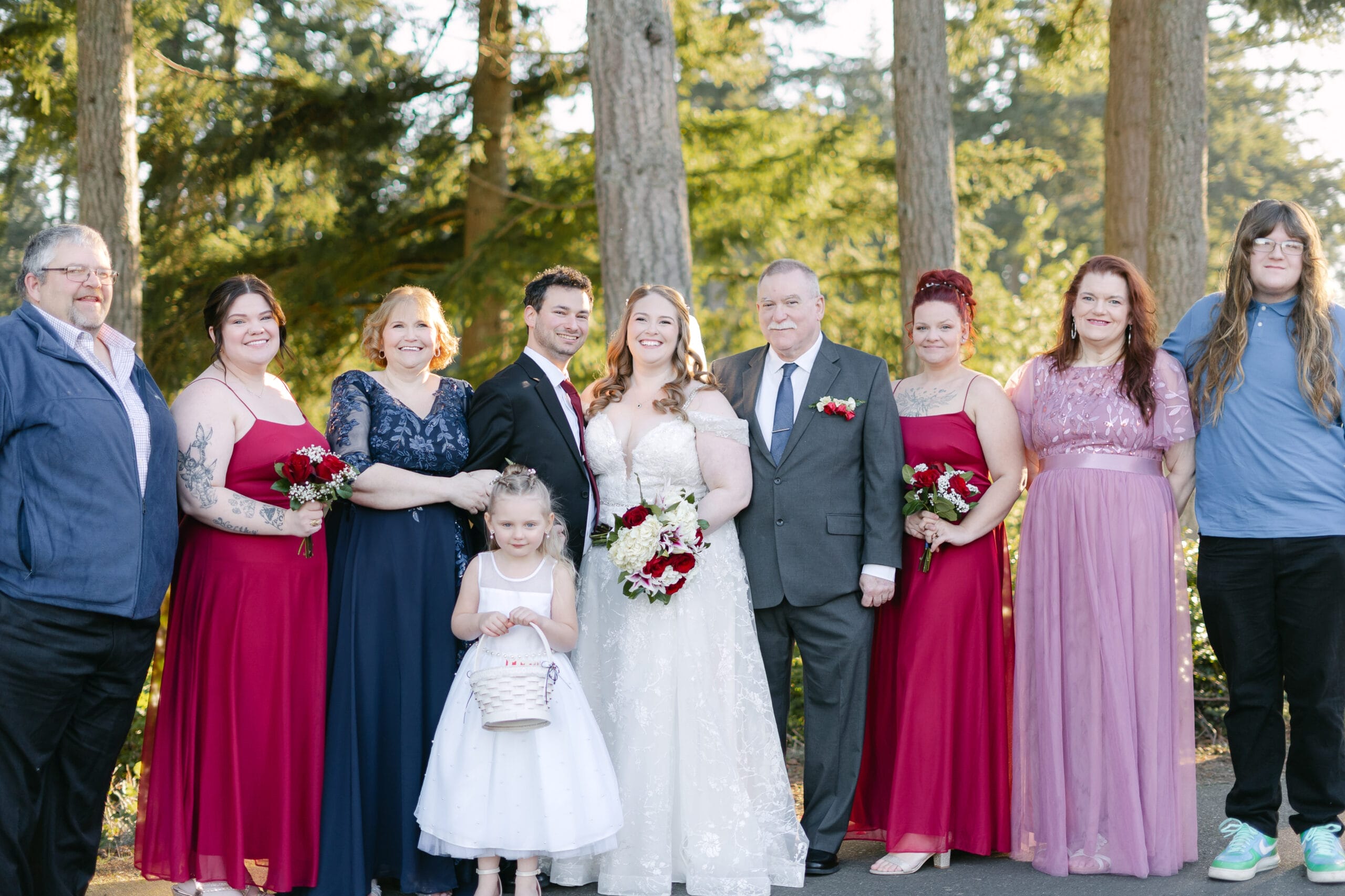 Bride and groom with their wedding party posing outdoors among trees, wearing formal attire and colorful dresses in burgundy, pink, and navy.