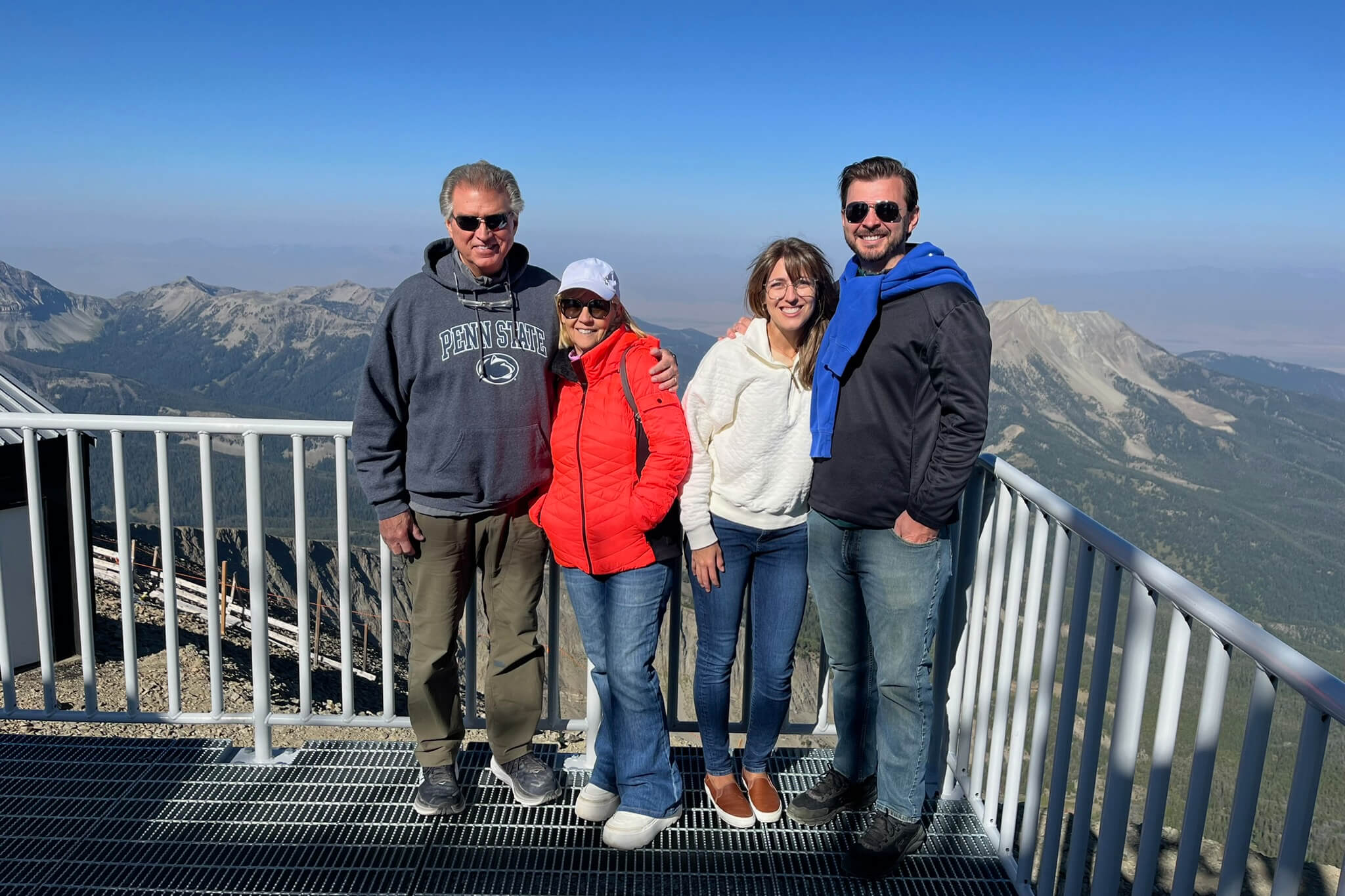 Four friends posing and smiling on a metal viewing platform with a mountain range in the background under a clear blue sky.
