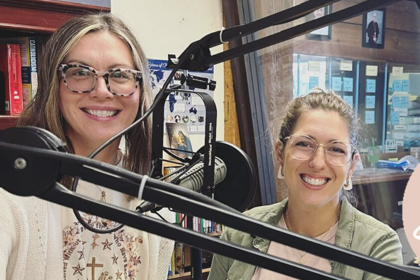Two women in a recording studio smiling behind large broadcast microphones.