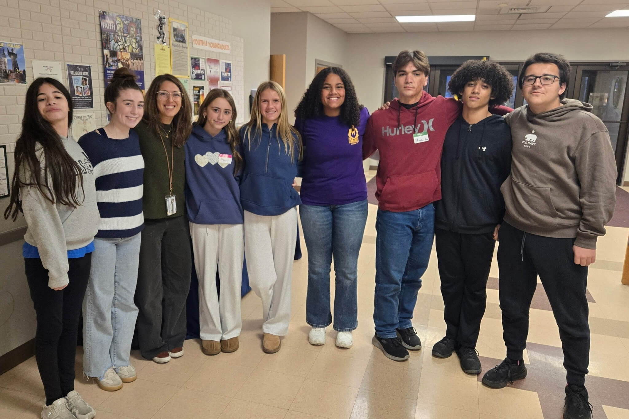 Group of students posing together in a school hallway, smiling at the camera with posters on the wall behind them.