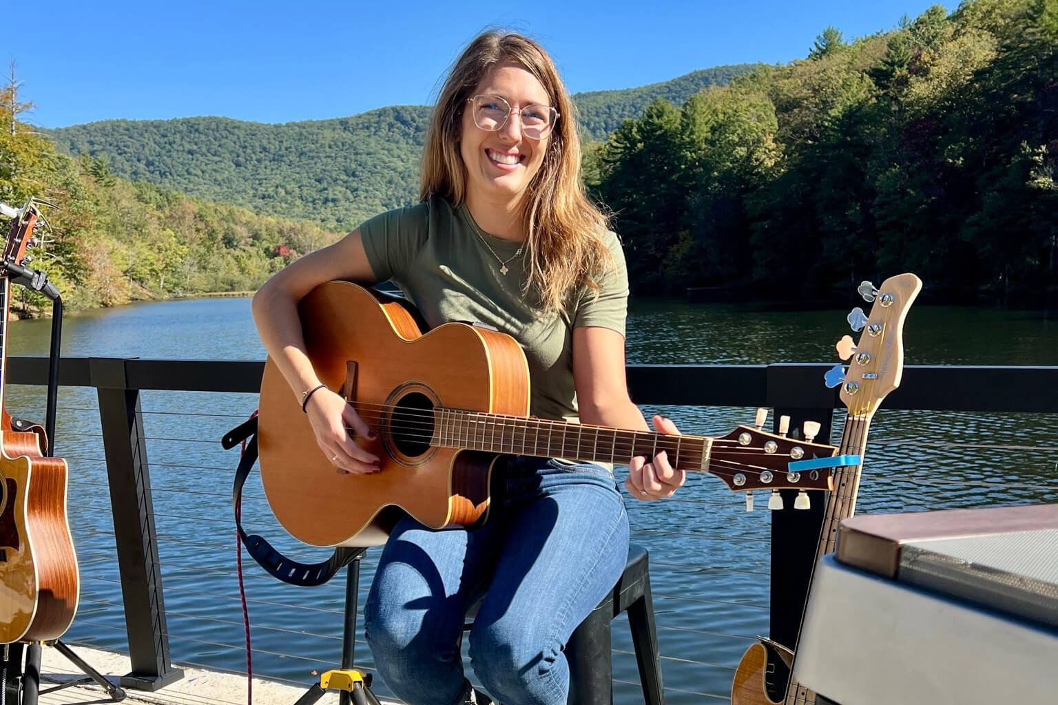 Smiling woman with glasses sits on a deck by a lake, playing an acoustic guitar with forested hills in the background.