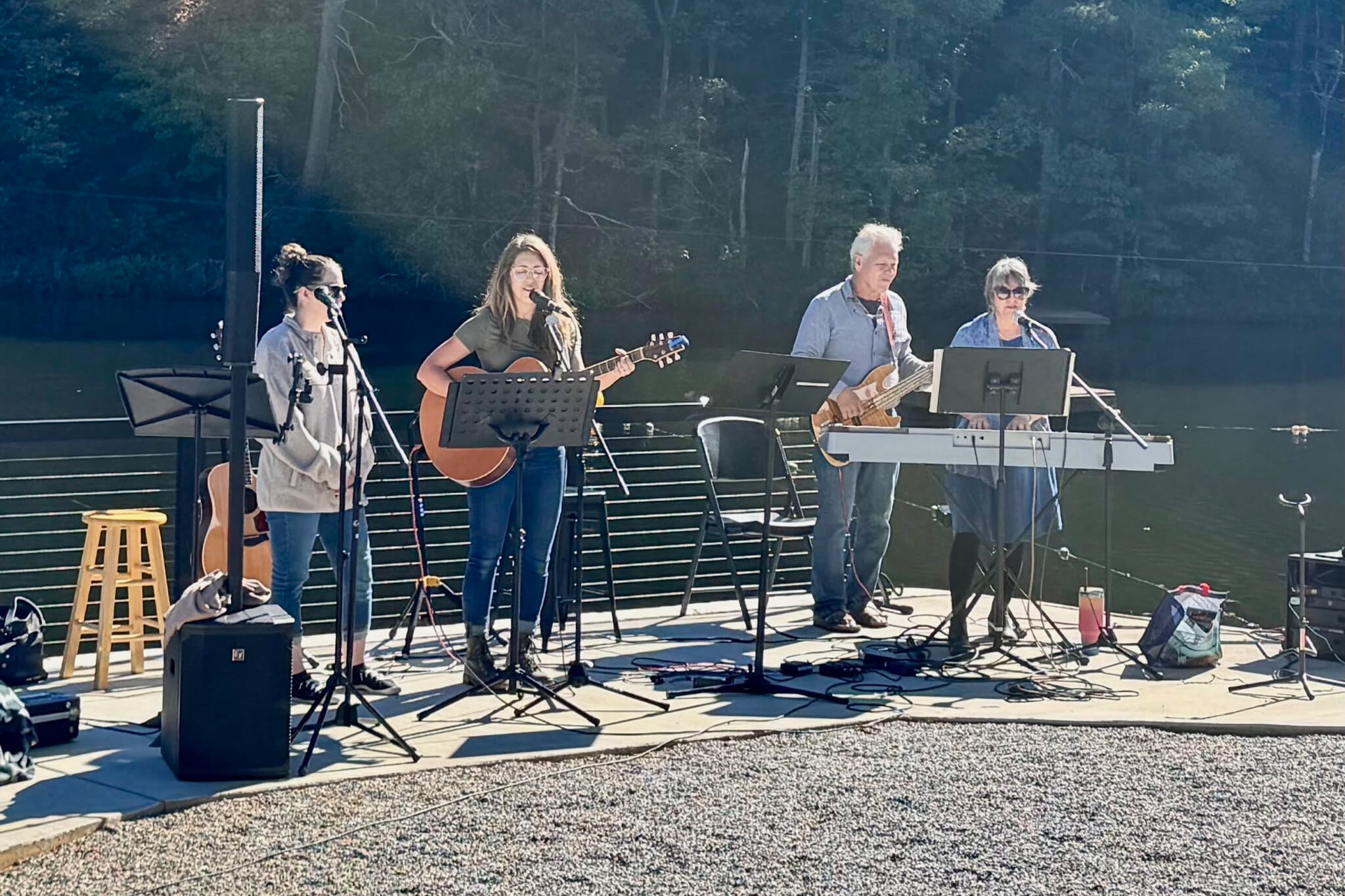 Four musicians perform on an outdoor dock by a lake, with guitars, keyboard, and microphones visible under a sunny sky.