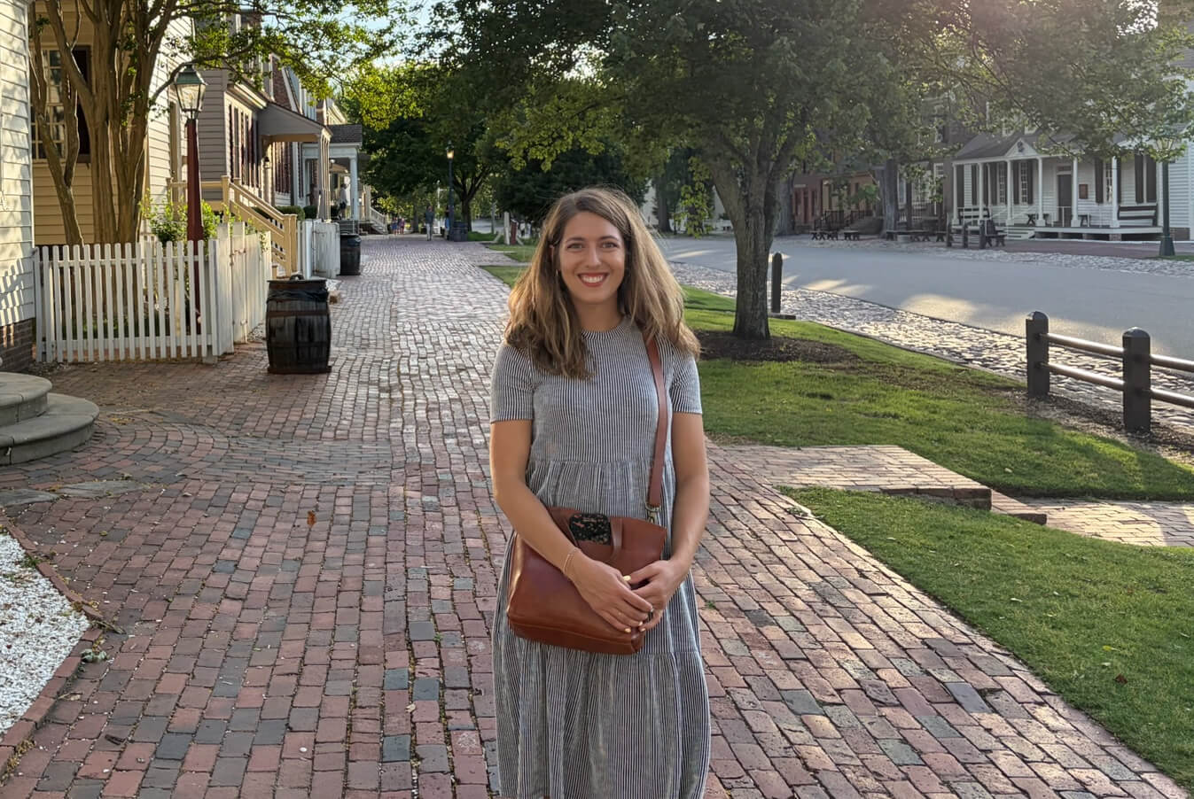 Smiling woman in a gray striped dress with a brown crossbody bag standing on a brick sidewalk along a tree-lined, historic street with houses nearby.