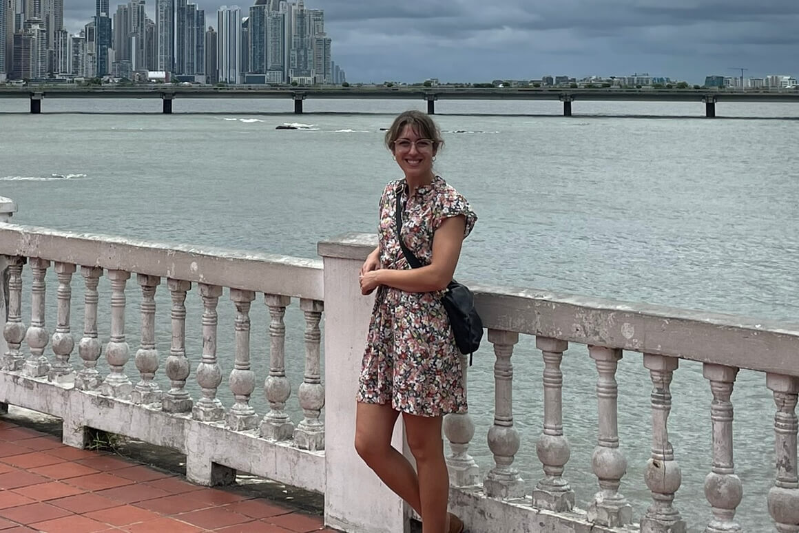 Smiling woman in a floral dress stands by a railing along the river, with a city skyline and bridge in the background.