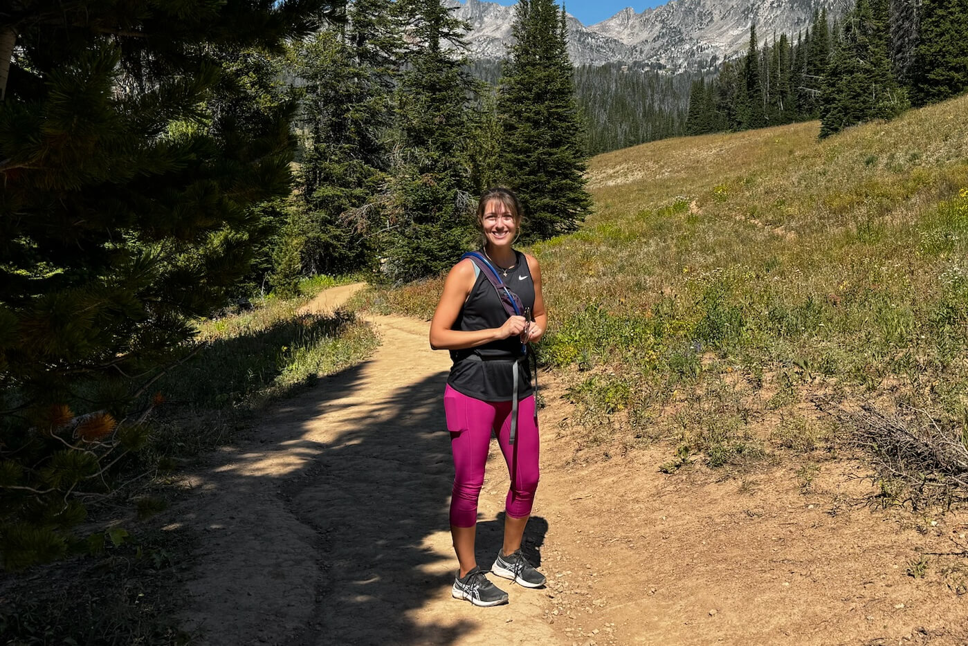 Woman hiking on a dirt trail in a mountain meadow, wearing a black tank top, magenta leggings and running shoes, smiling at the camera with mountains in the background.
