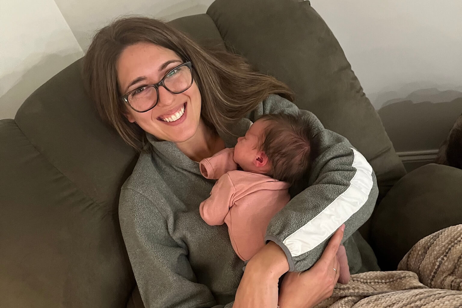 Woman with glasses smiling while holding a newborn in a pink outfit on a gray couch.