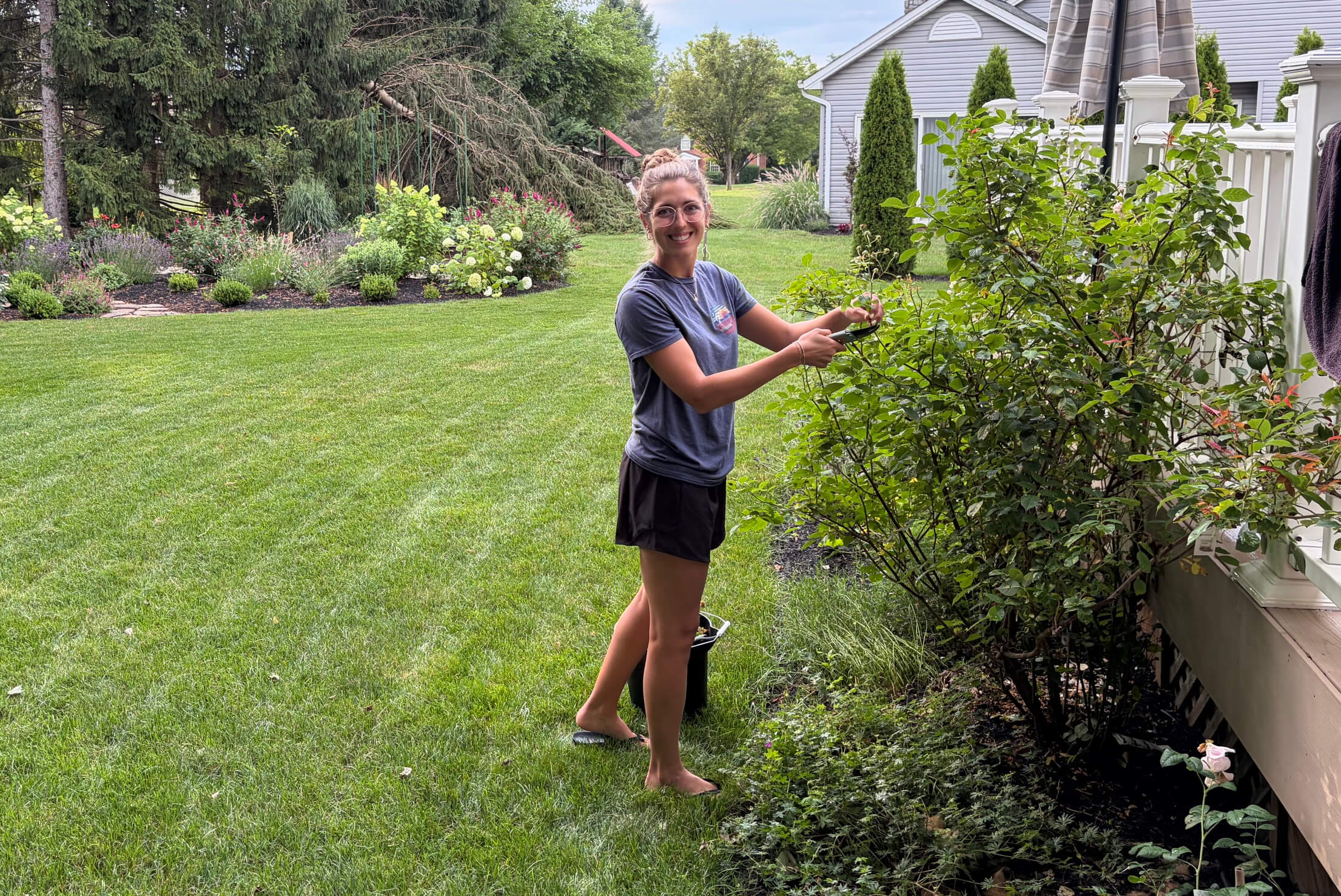 Smiling woman in a gray T-shirt stands barefoot on a lawn, pruning a shrub with shears beside a white fence and patio umbrella.