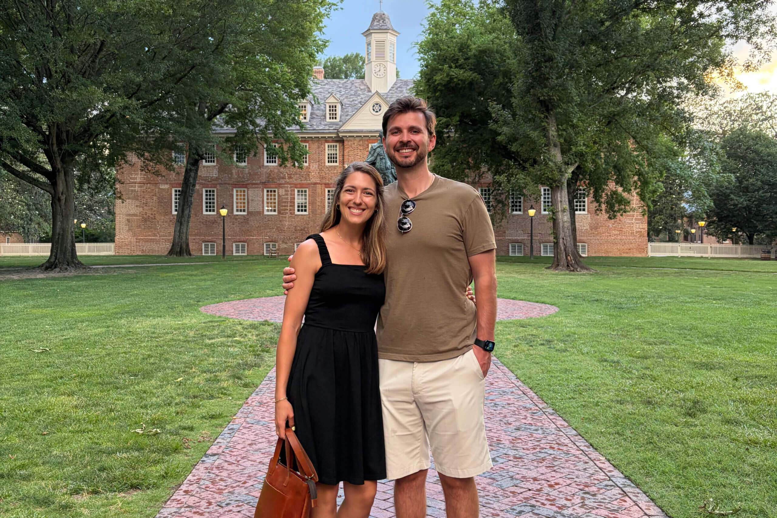 Smiling couple standing on a brick path in a park, with a red brick campus building and clock tower behind them.