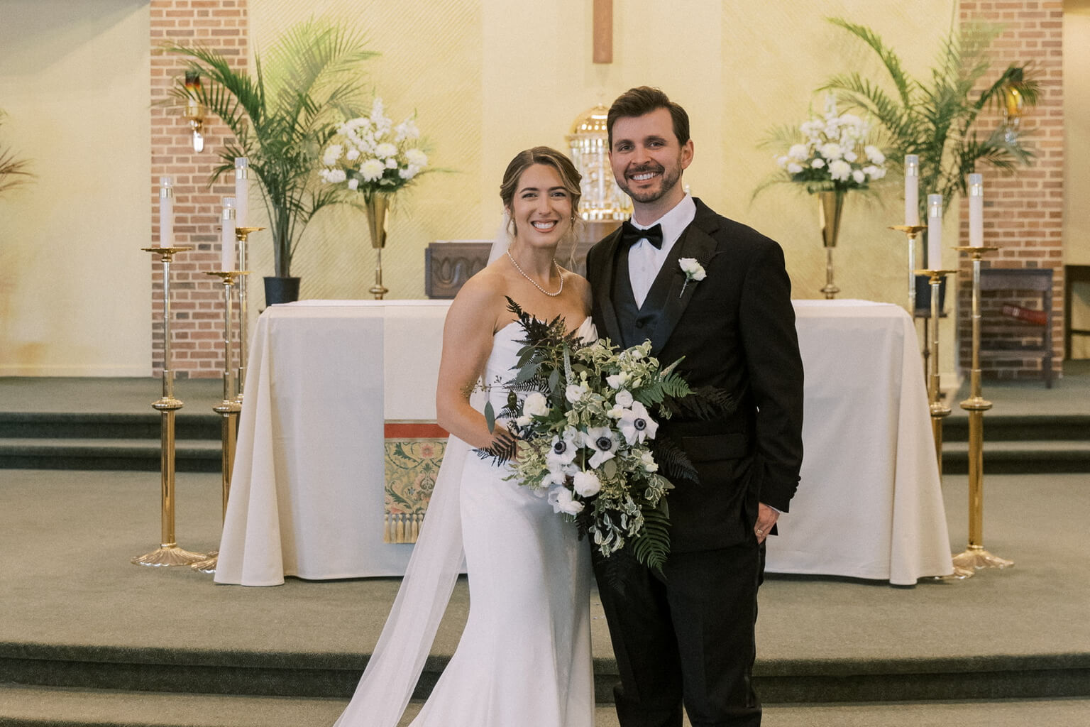 Bride in a white gown holds a white-flower bouquet and smiles beside her groom at the church altar.