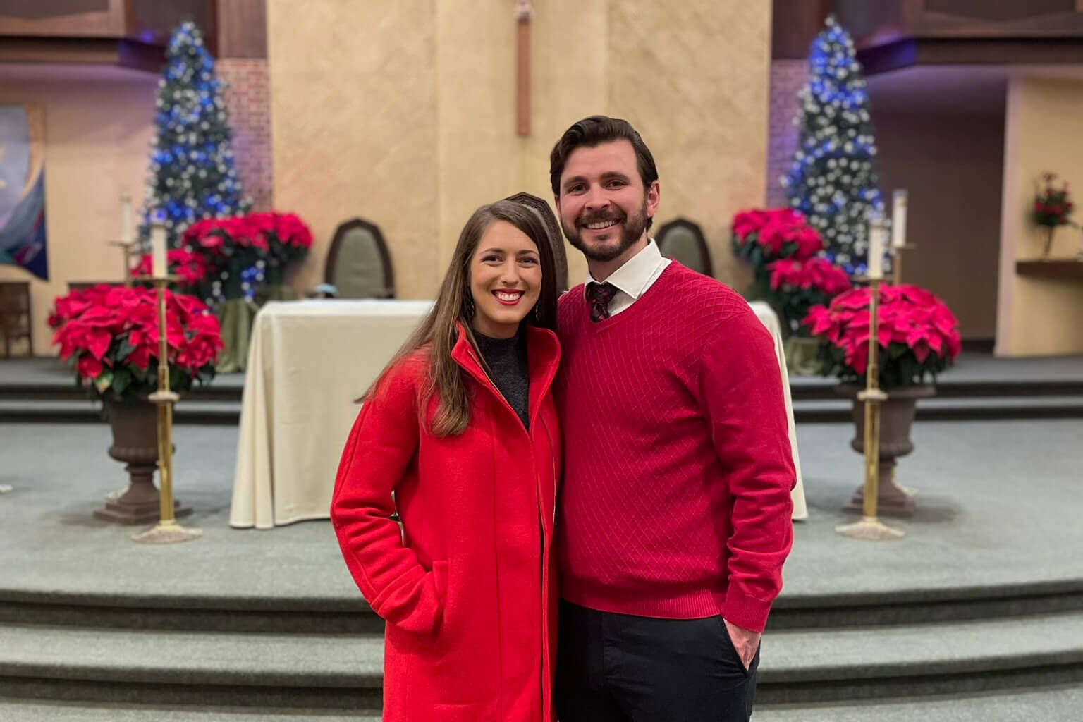 Smiling couple in red outfits posing in a church sanctuary decorated with poinsettias and Christmas trees.