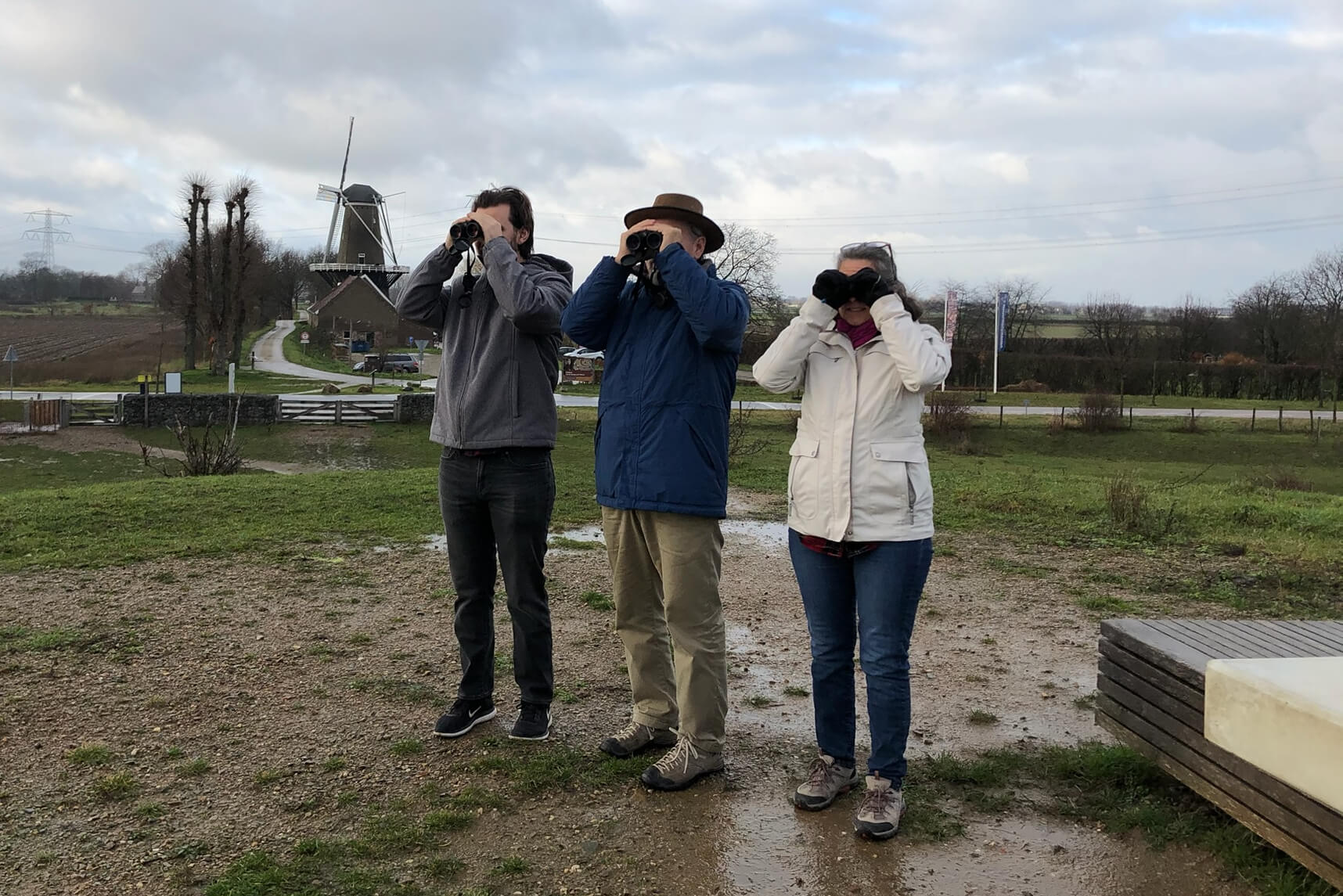Three adults stand in a damp, rural area looking through binoculars at the distance, with a windmill and fields in the background.