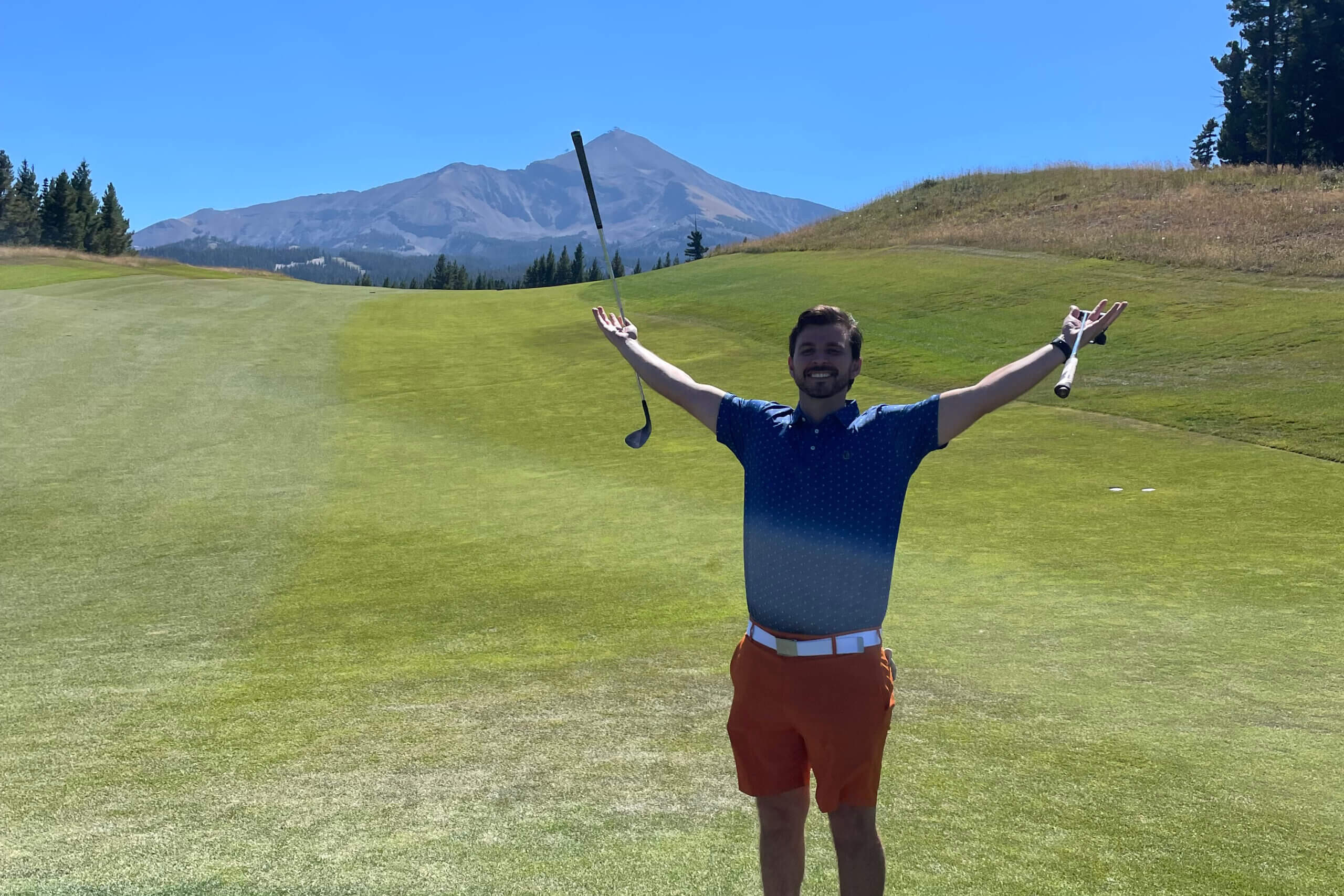Man on a golf course arms raised with two clubs, celebrating, with a mountain range in the distance.